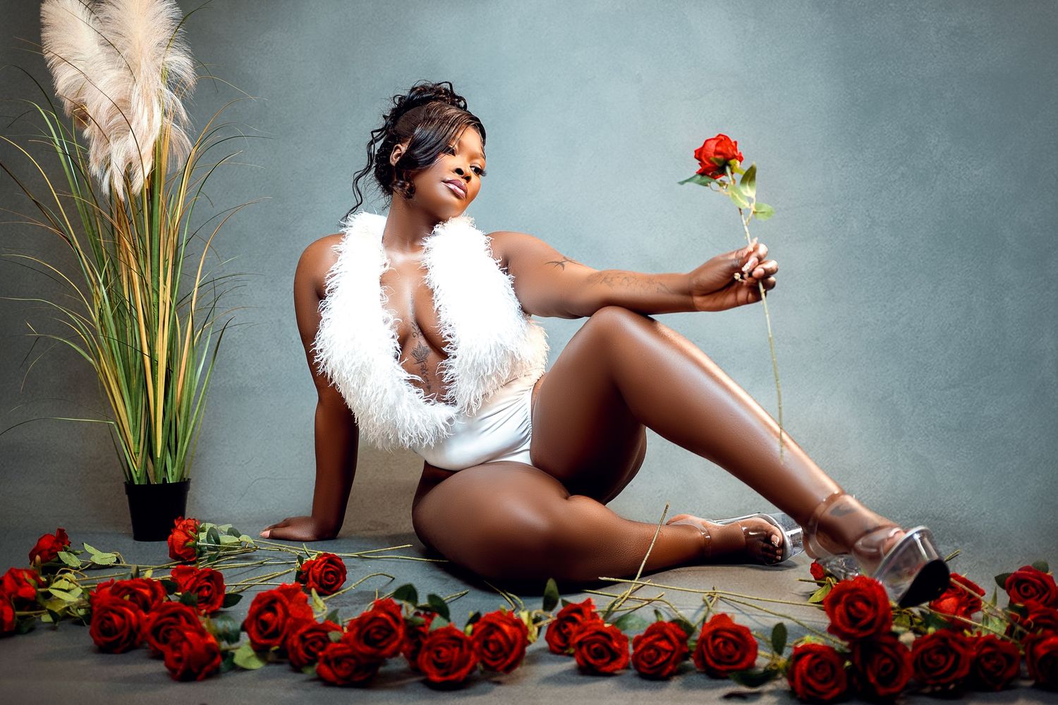 Woman in white feather bodysuit sitting on floor holding red rose, surrounded by roses, in professional studio setting.