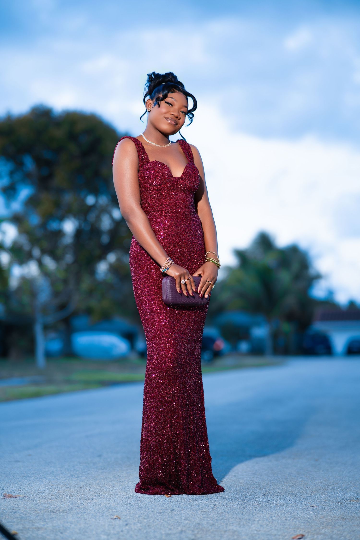 Young woman in elegant burgundy sequin gown holding clutch purse poses on suburban street at dusk.