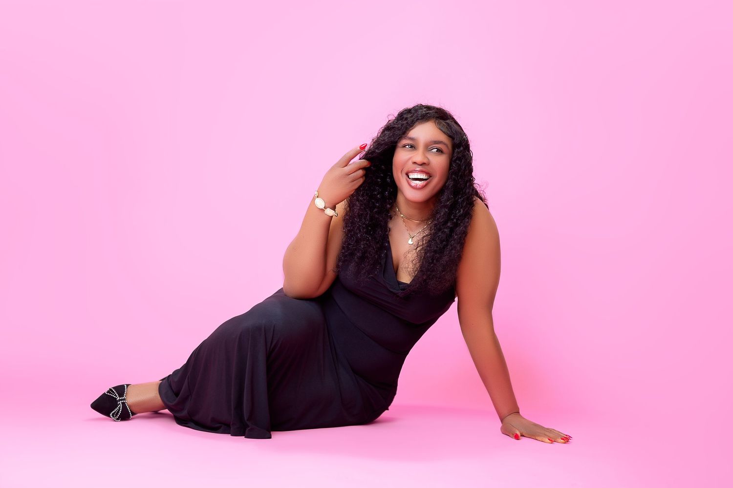Woman in black jumpsuit sitting on pink background, laughing joyfully with hand in hair.