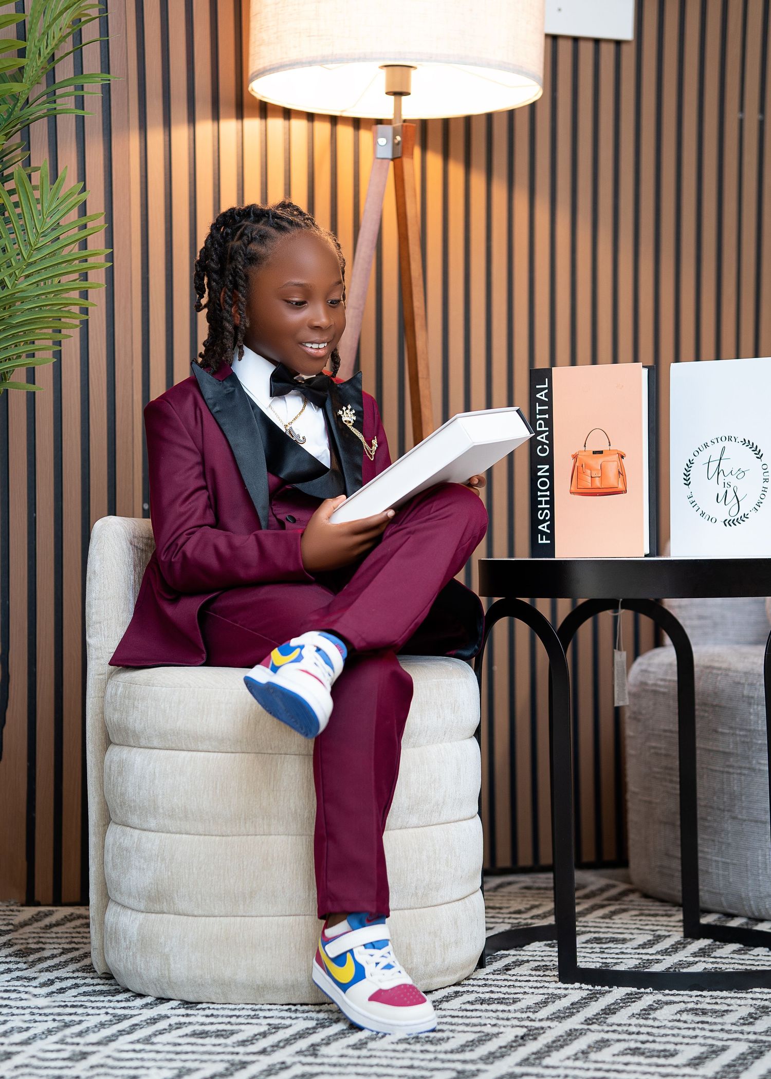 Young child in burgundy tuxedo reads book on white chair beside Fashion Capital display with Nike sneakers.