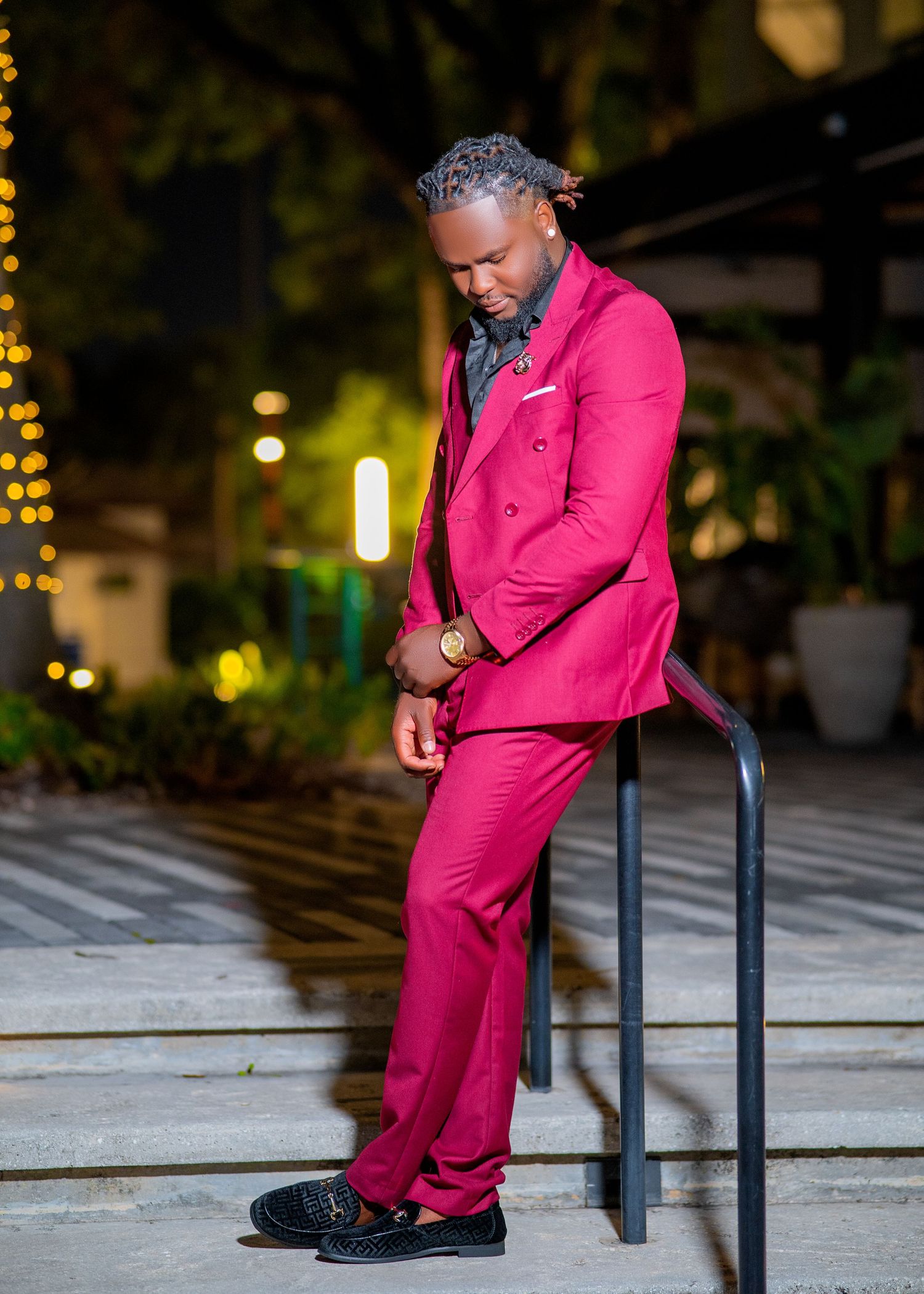 Man in a bold magenta double-breasted suit leaning on a railing outdoors at night with bokeh lights in background.