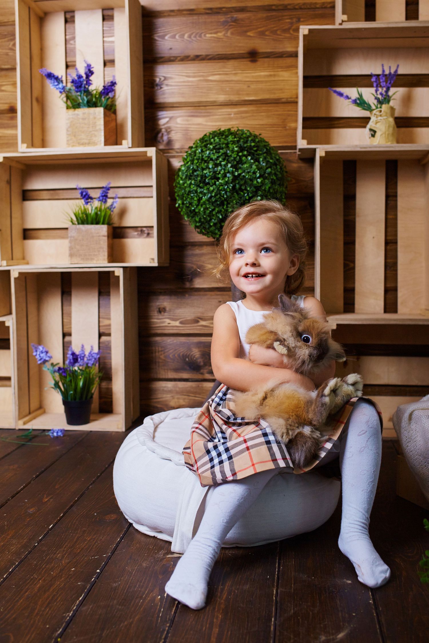 Child in white outfit cuddles with small Pomeranian dog against rustic wooden wall with shelving.