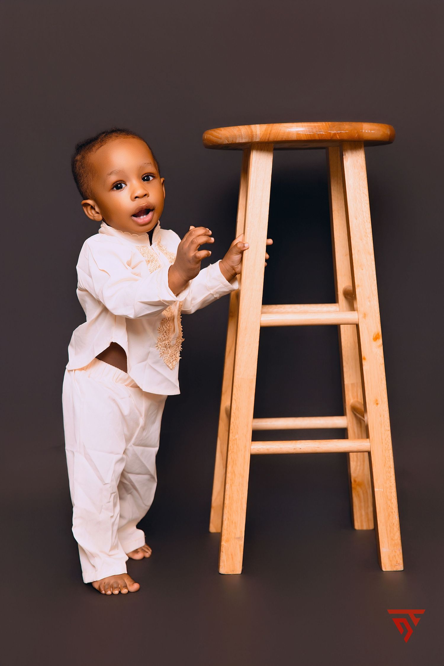 Baby in white outfit crawling near wooden stool against dark background.