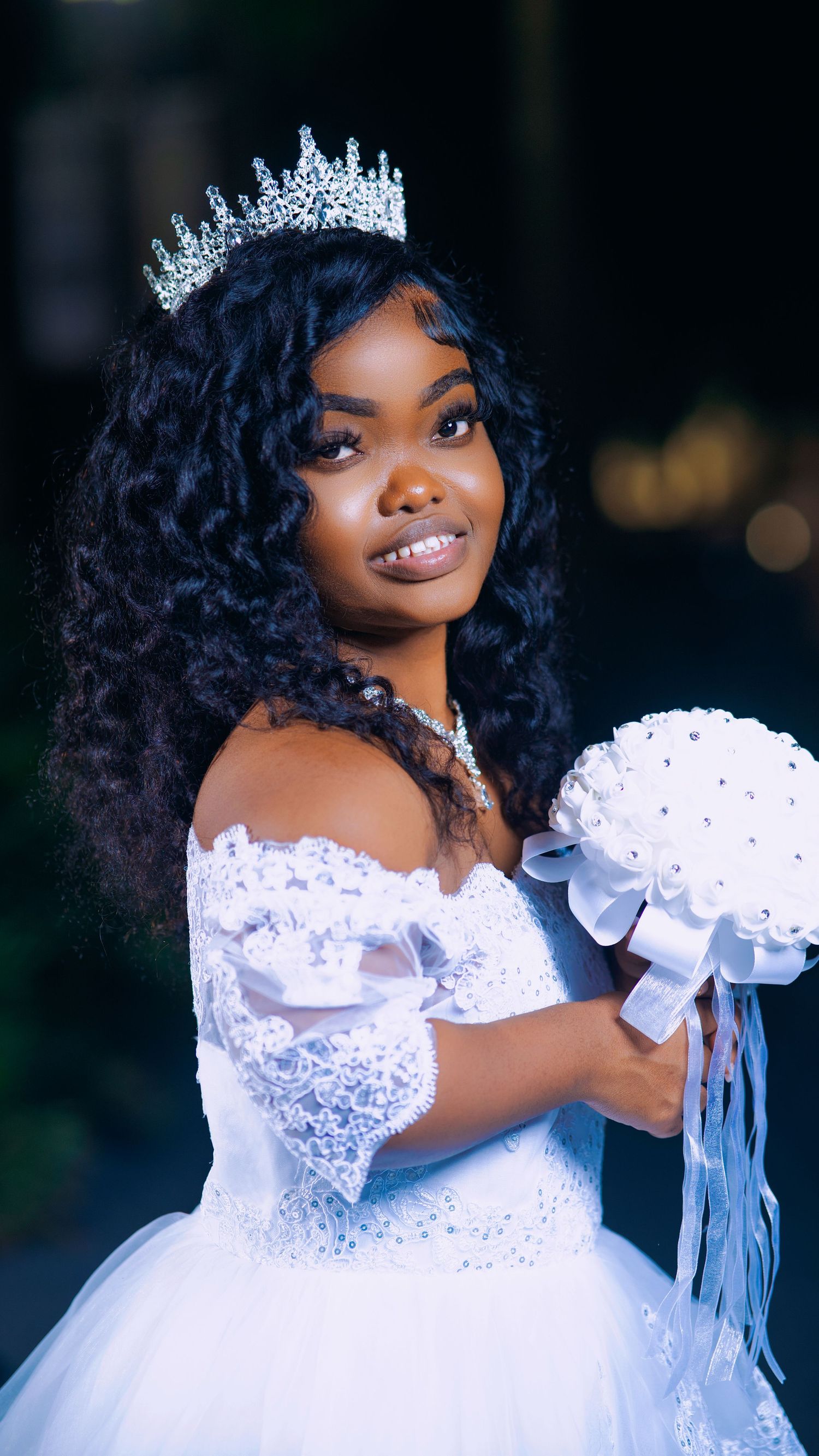 Bride in sparkling tiara and off-shoulder white gown holds decorative bouquet against dark background.