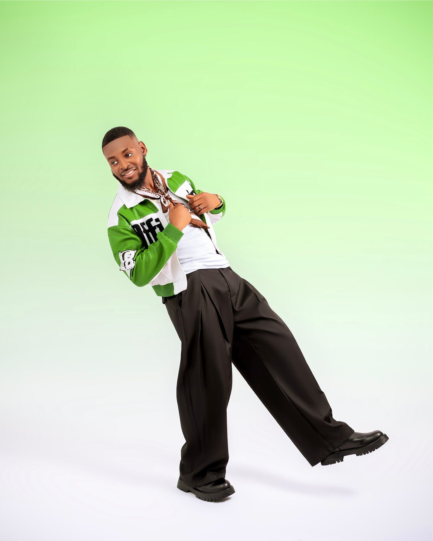 Young man posing confidently in green and white varsity jacket with wide-leg black trousers against green background.