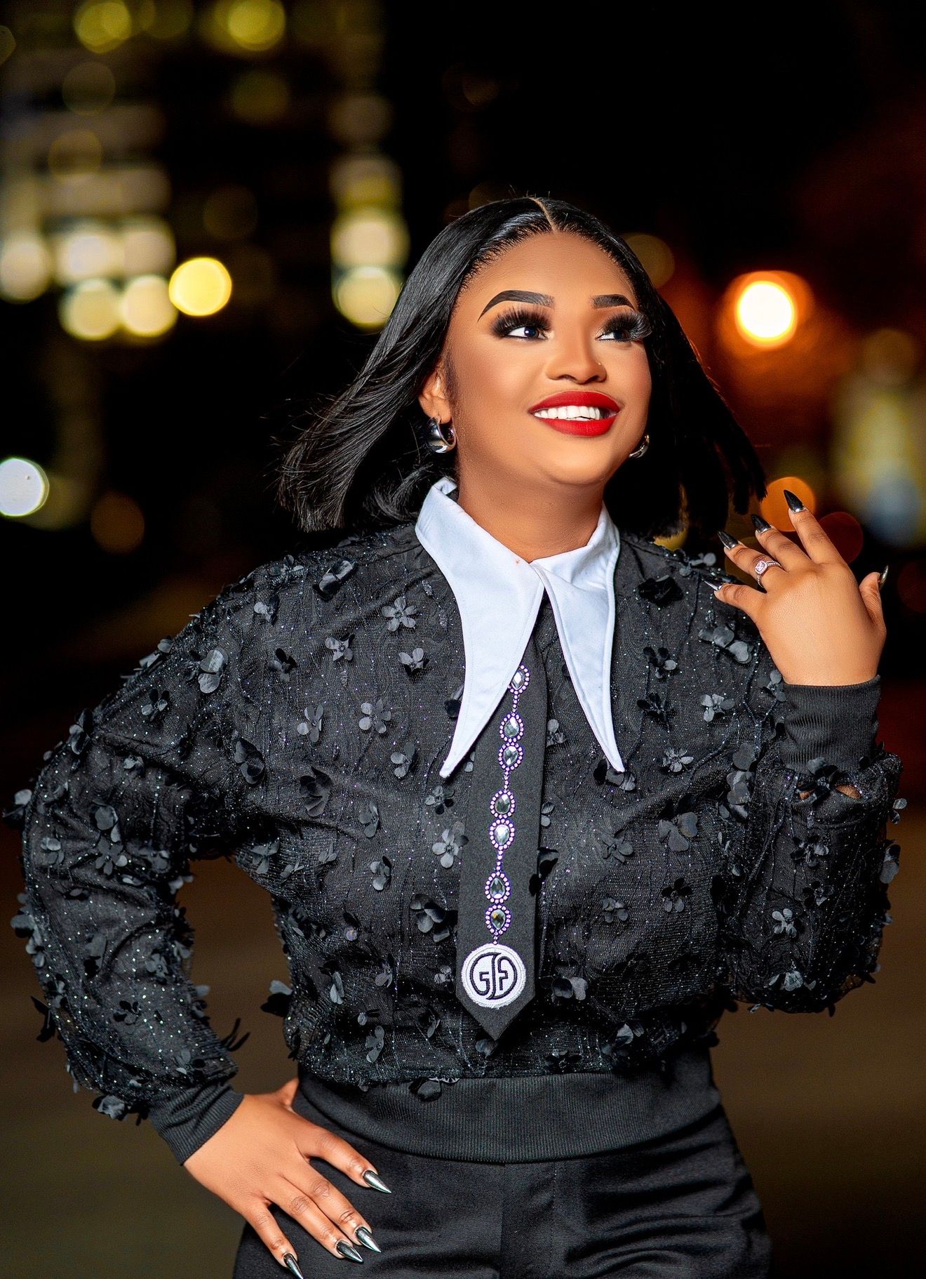 Woman in black floral embroidered top with white collar and decorative tie, posing confidently at night.
