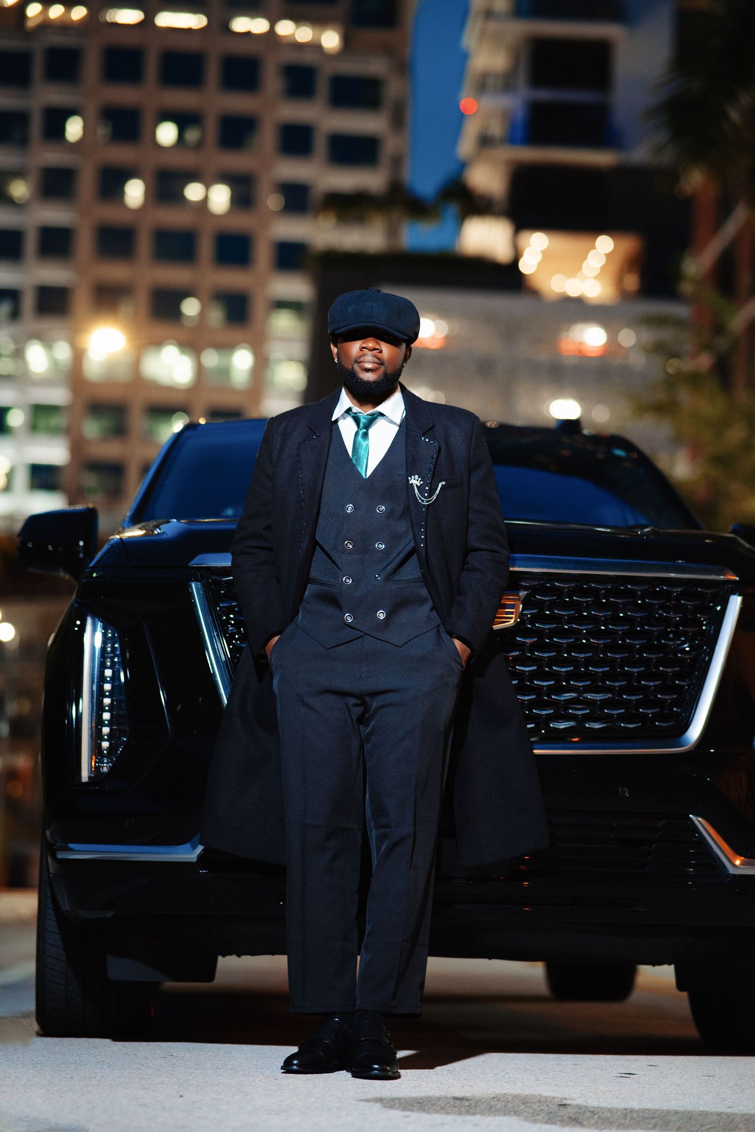 Stylish man in dark suit, vest, and newsboy cap poses in front of black SUV with city lights at night.