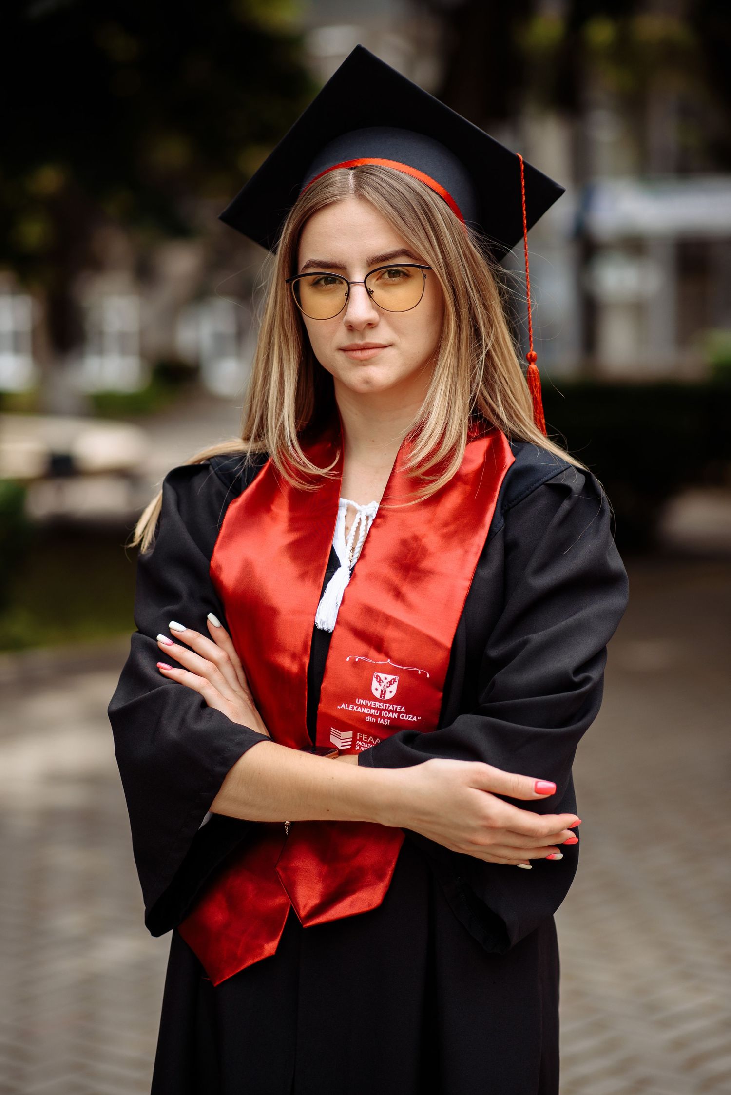 Graduate in black cap and gown with red stole poses outdoors on campus.