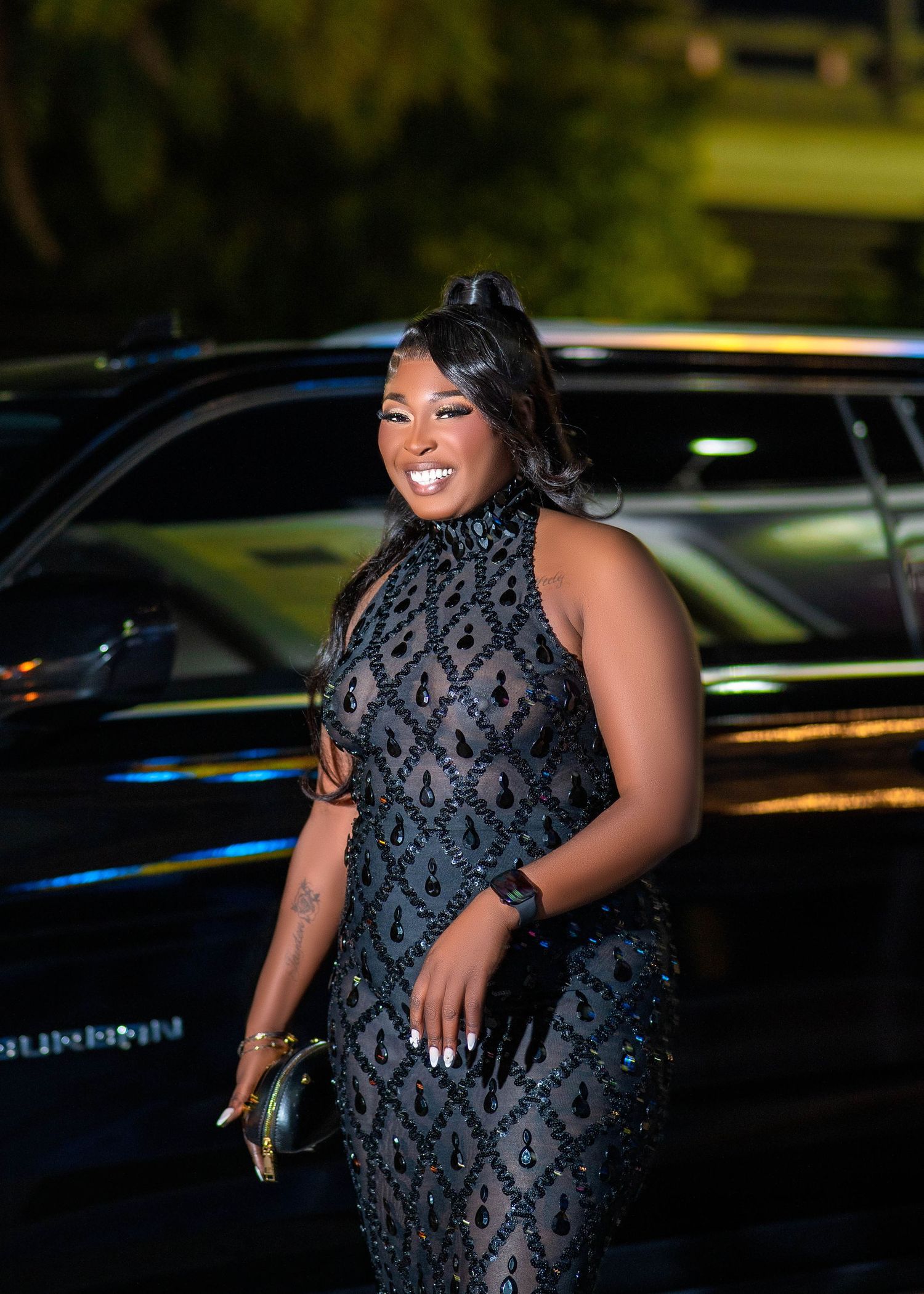 A person wearing a black sequined dress poses confidently against a backdrop of parked cars in an evening setting.