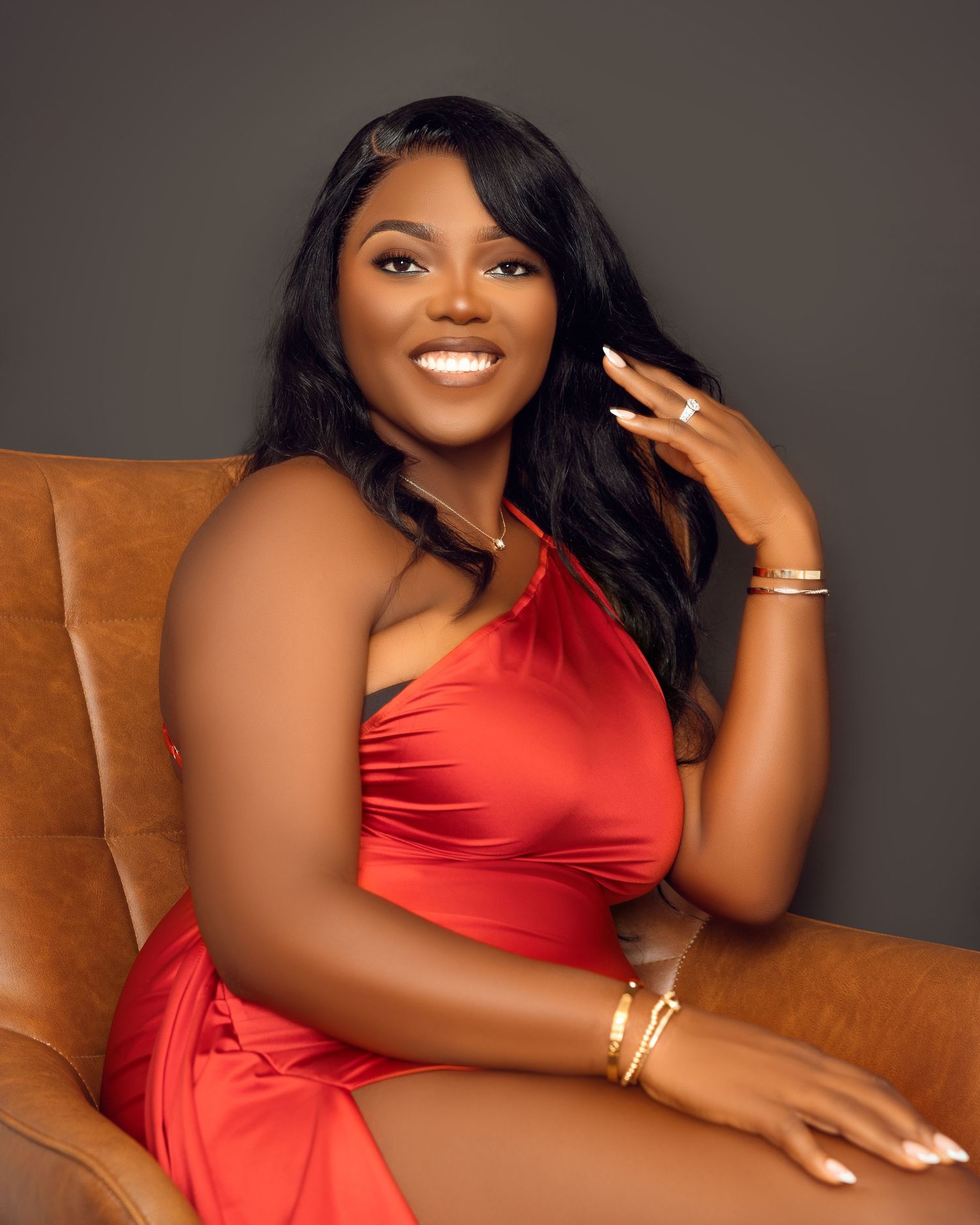Woman in red dress poses confidently on brown leather chair against dark background.