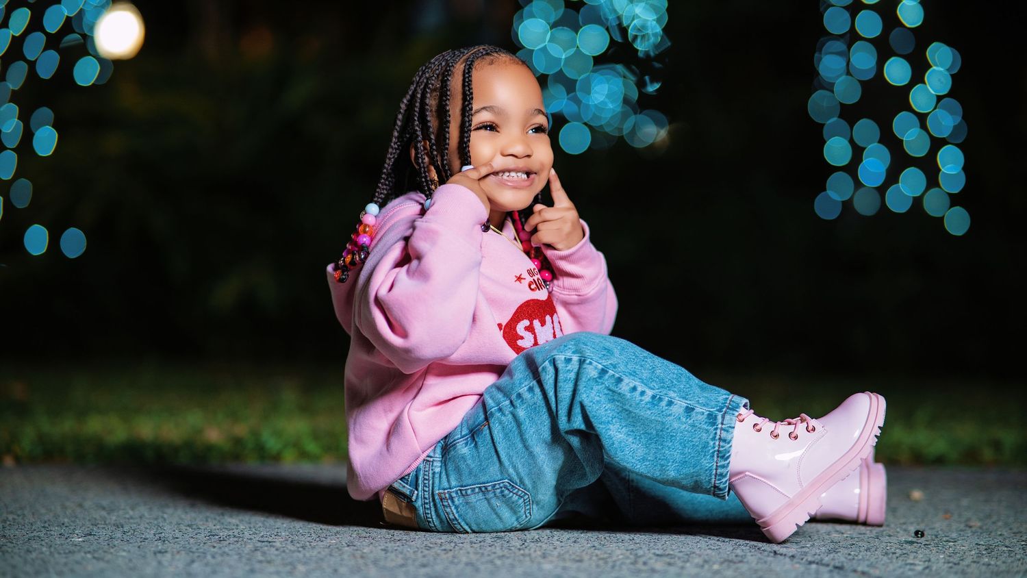 Child in pink sweater and jeans plays on concrete path with blue lights in background.