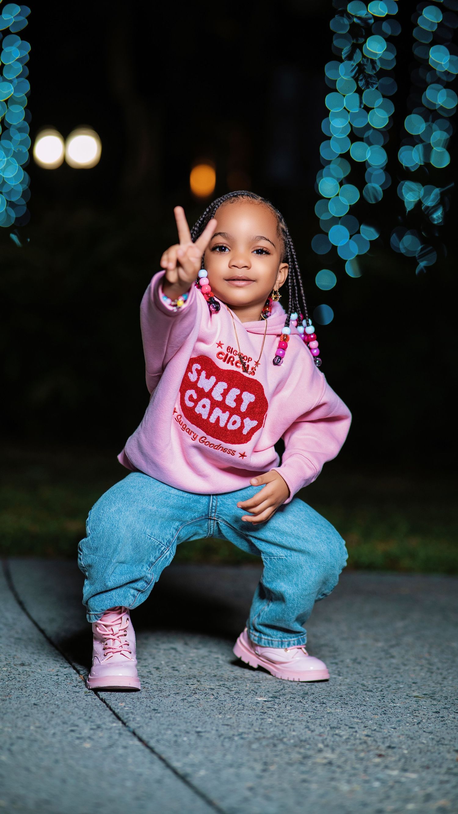 Toddler in pink sweater and jeans crouches on sidewalk with blue bokeh lights in background.