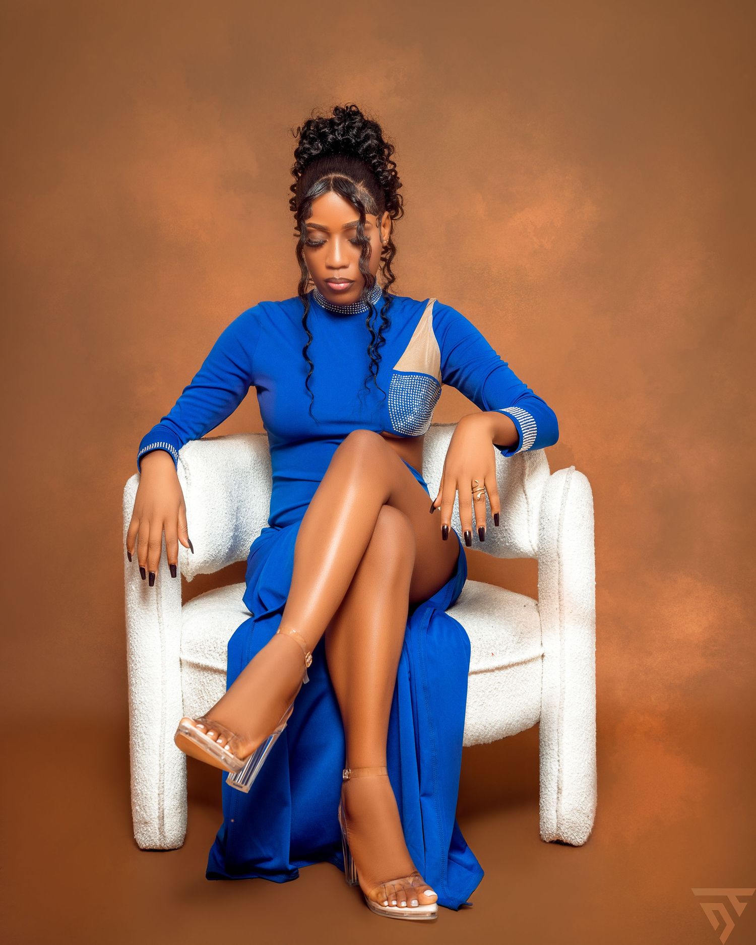 Fashion portrait series in royal blue dress seated on white armchair against warm brown background.