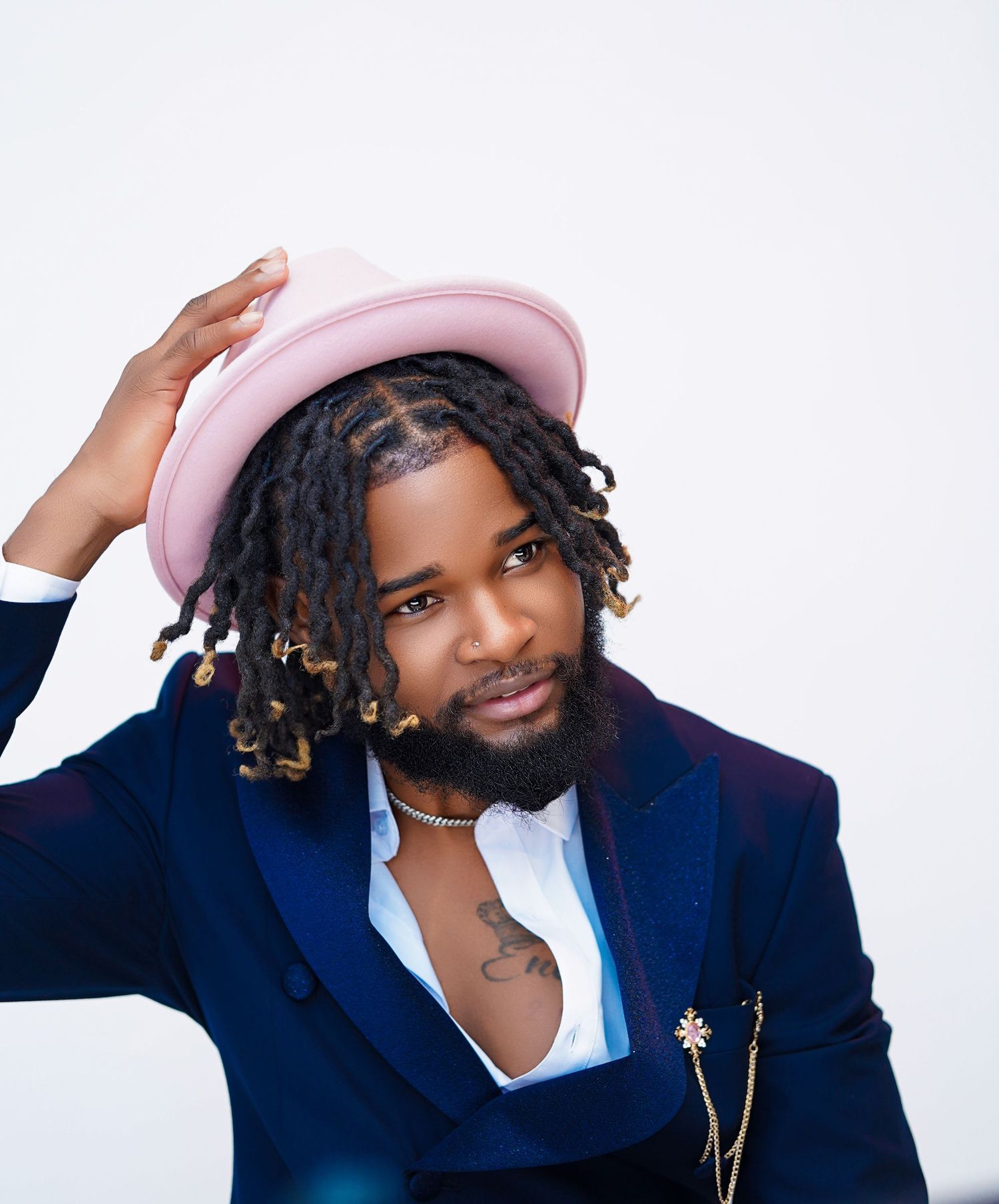 Stylish man with dreadlocks wearing a pink hat and navy blazer poses confidently against a white background.