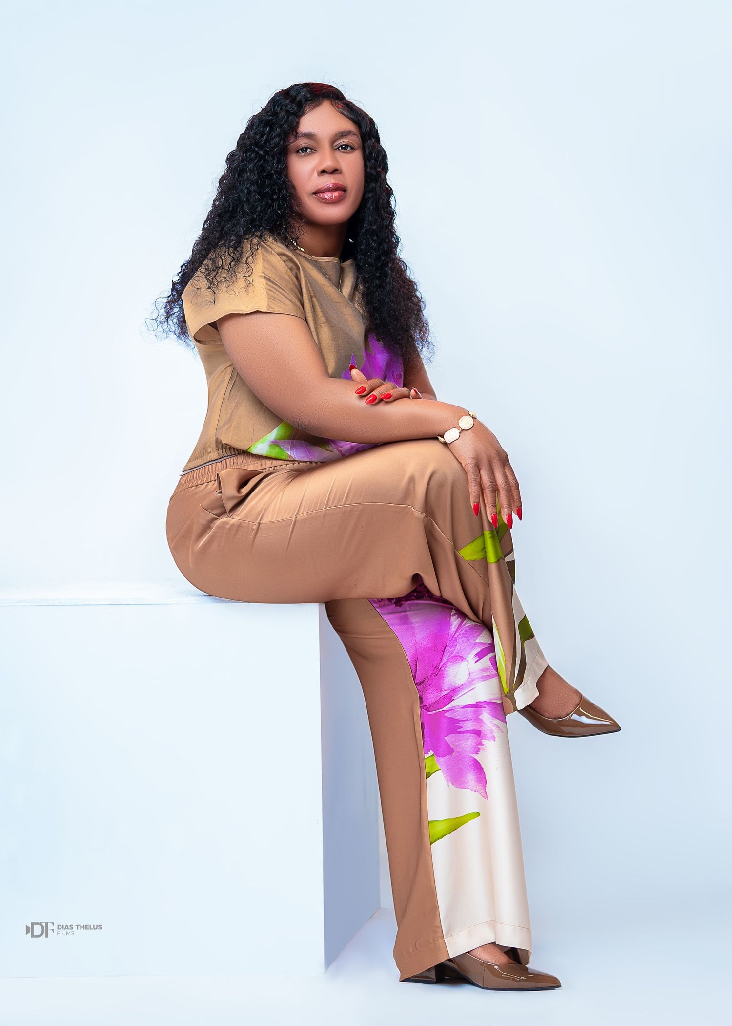 Woman in tan outfit with floral wide-leg pants sitting on white cube, posing confidently in a professional photo shoot.