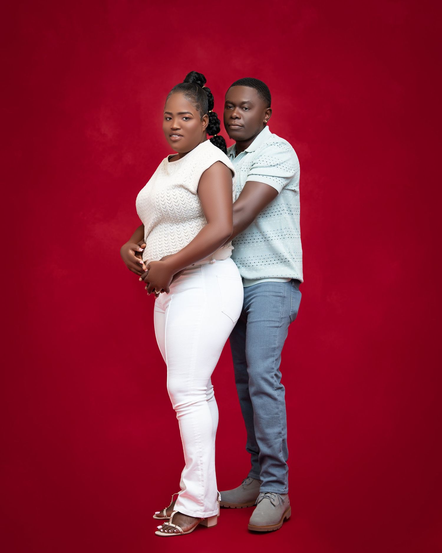 A couple poses against a red background, woman in white outfit and man in light blue shirt with jeans.