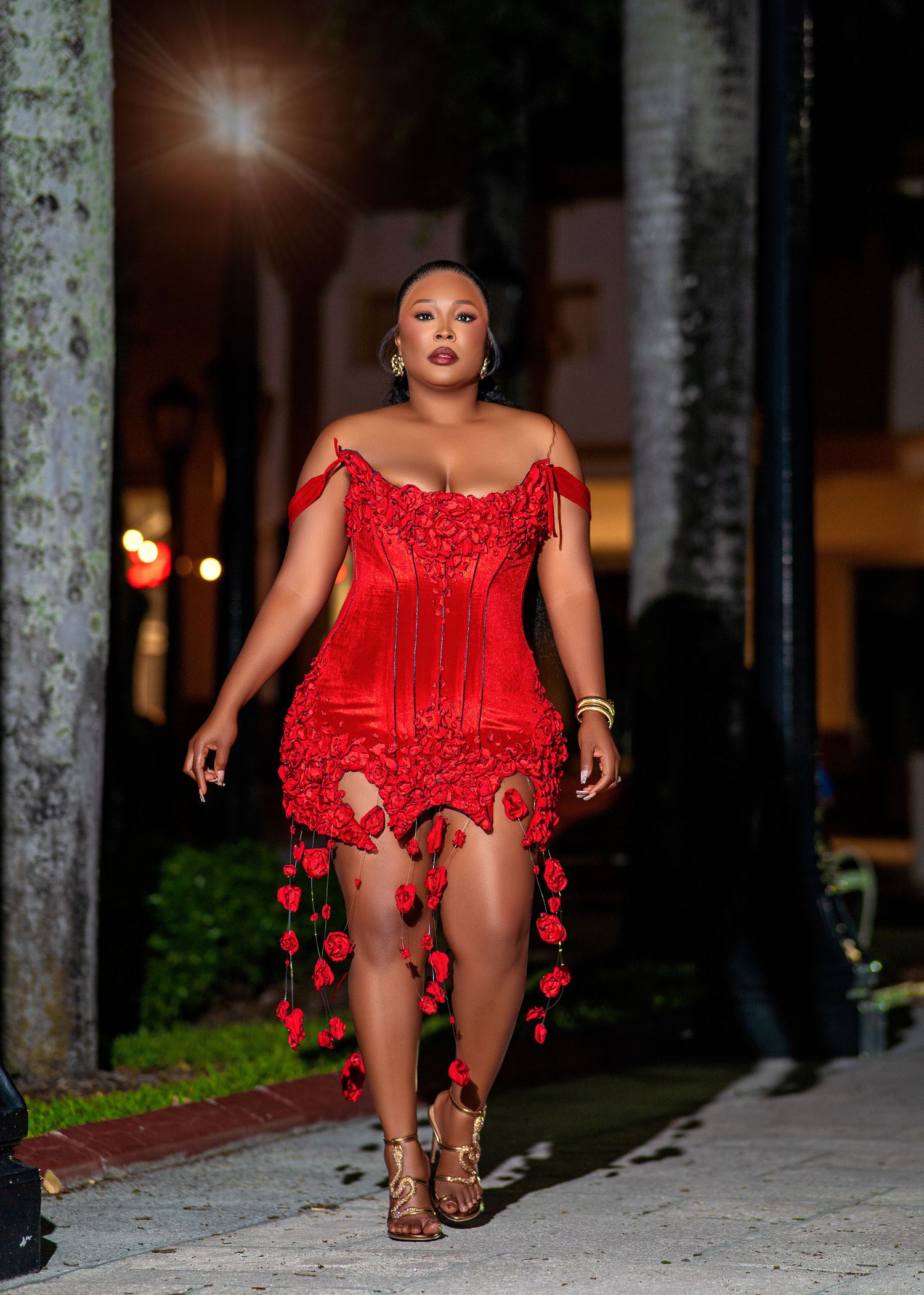 Woman in red off-shoulder mini dress with rose details walks confidently at night.