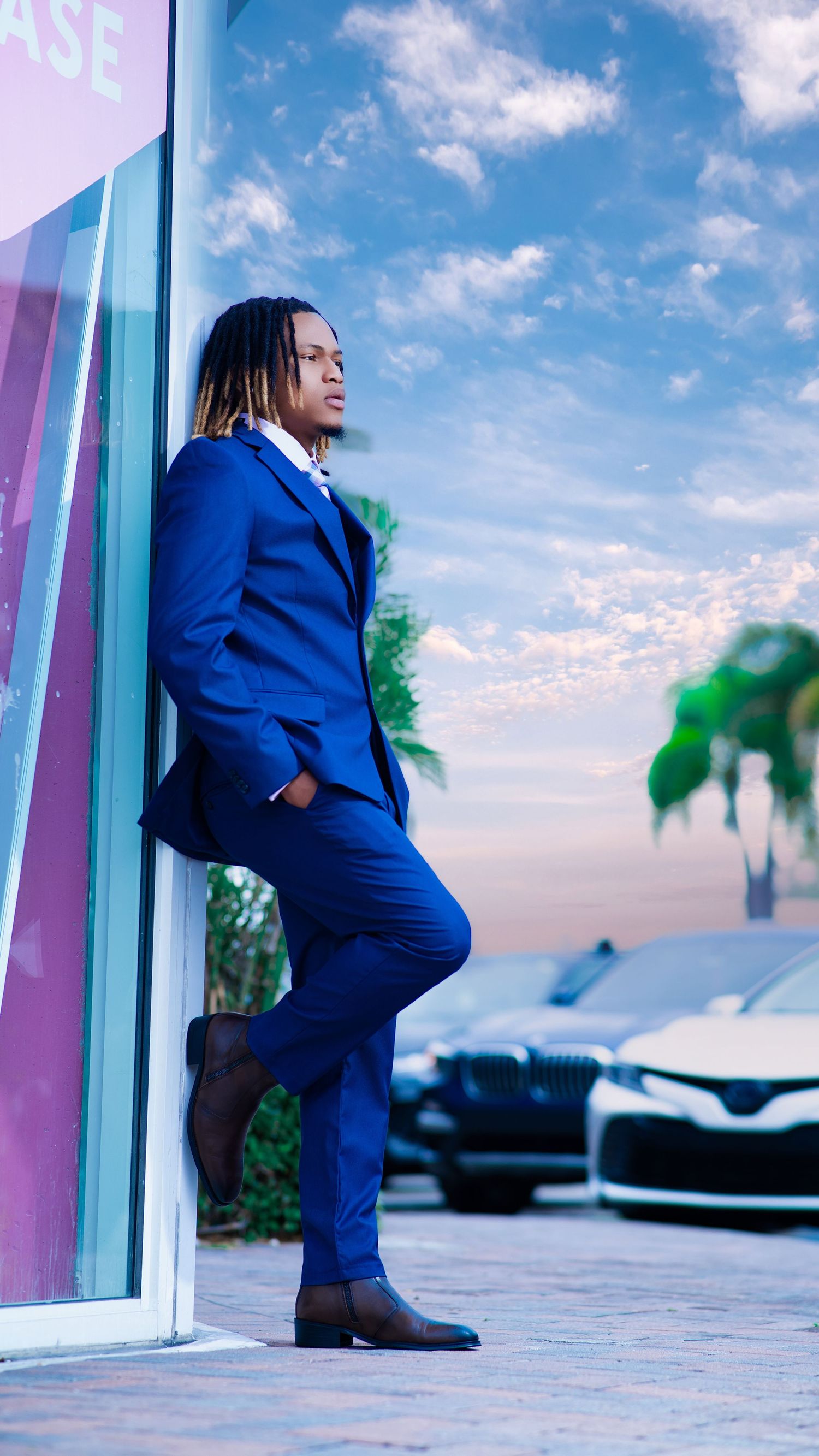 A stylish man in a royal blue suit with dreadlocks leans against a building near luxury cars under a dramatic sky.