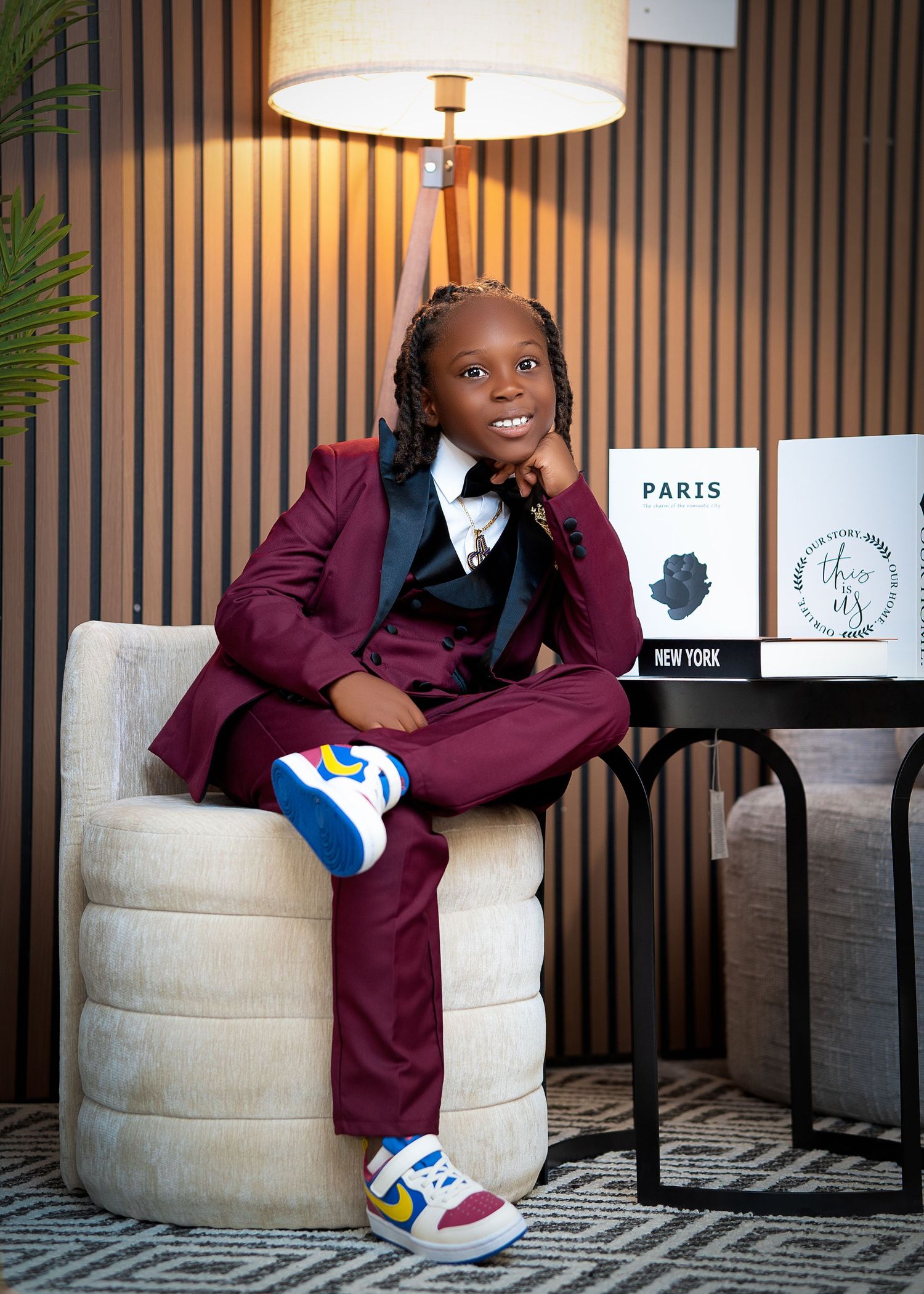 Young child in burgundy suit and colorful sneakers poses confidently in modern interior setting with Paris and New York prints.