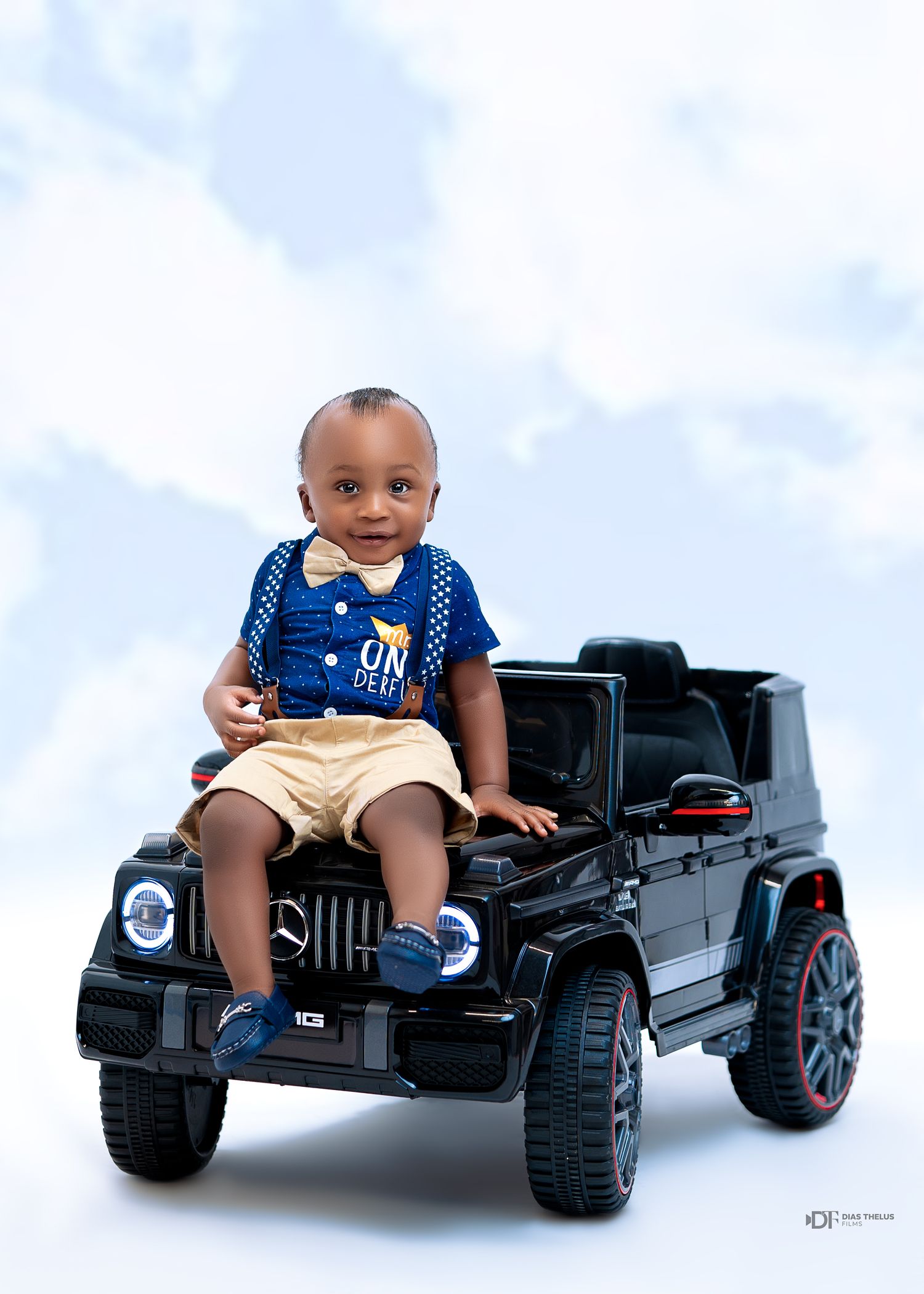 Smiling toddler boy in blue shirt and khaki shorts sitting on a black Mercedes toy ride-on SUV car.