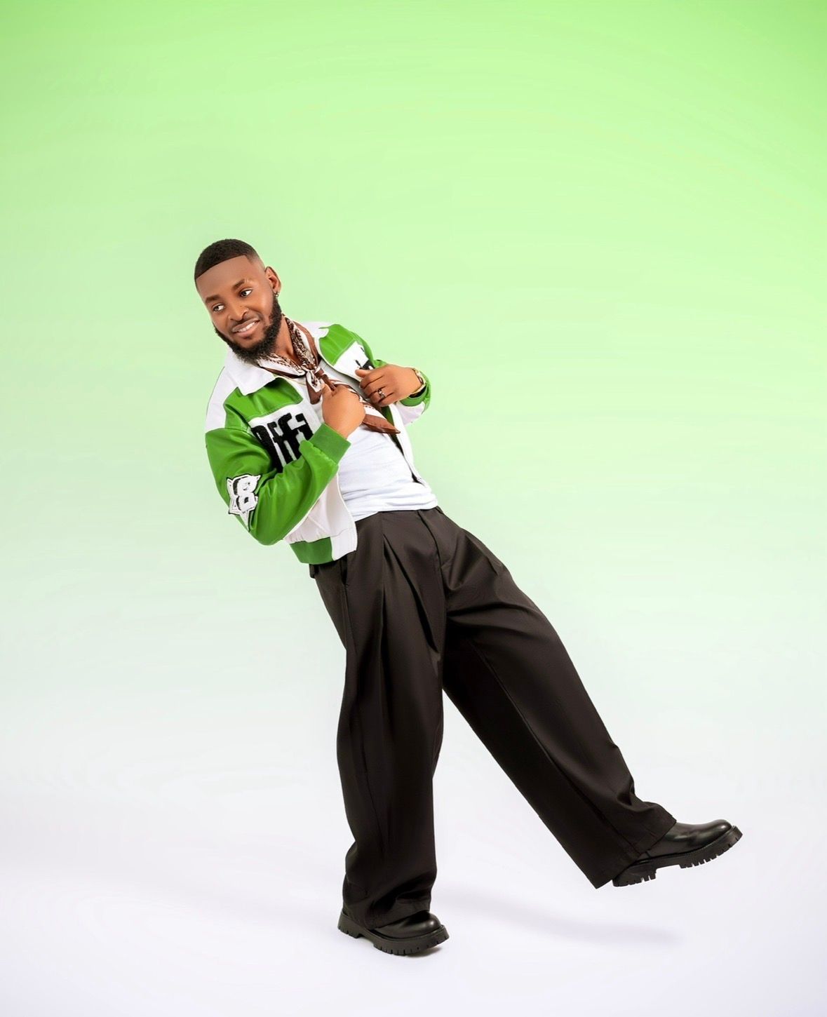 Young man posing confidently in green varsity jacket and wide-leg dark trousers against green gradient background.