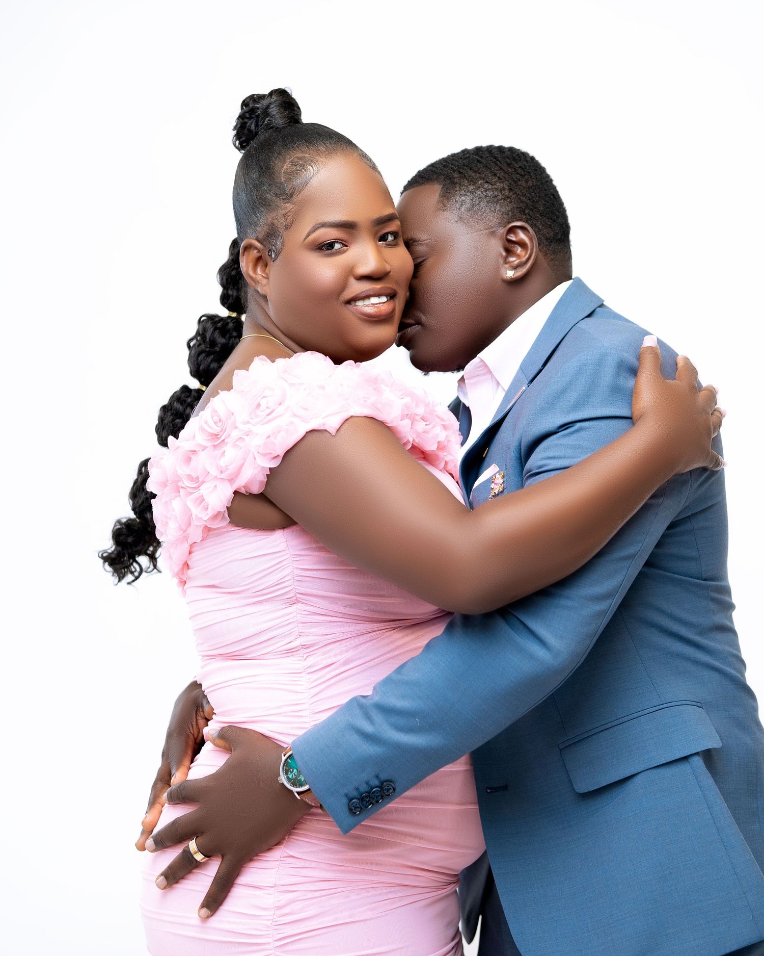 A couple shares a romantic embrace, man in blue suit kissing woman in pink dress against white background.