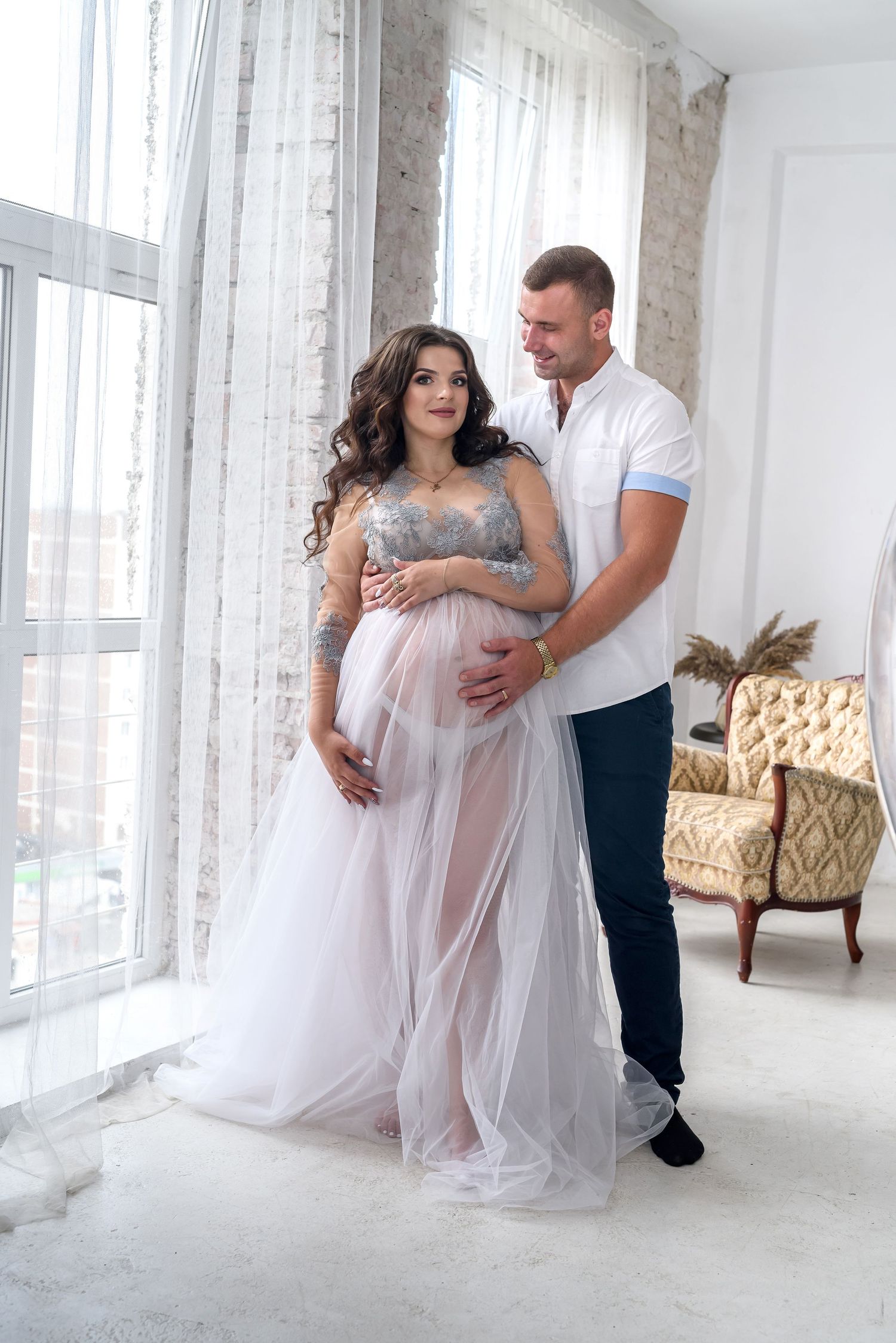 Romantic maternity photoshoot in white flowing dress with couple embracing in bright studio.