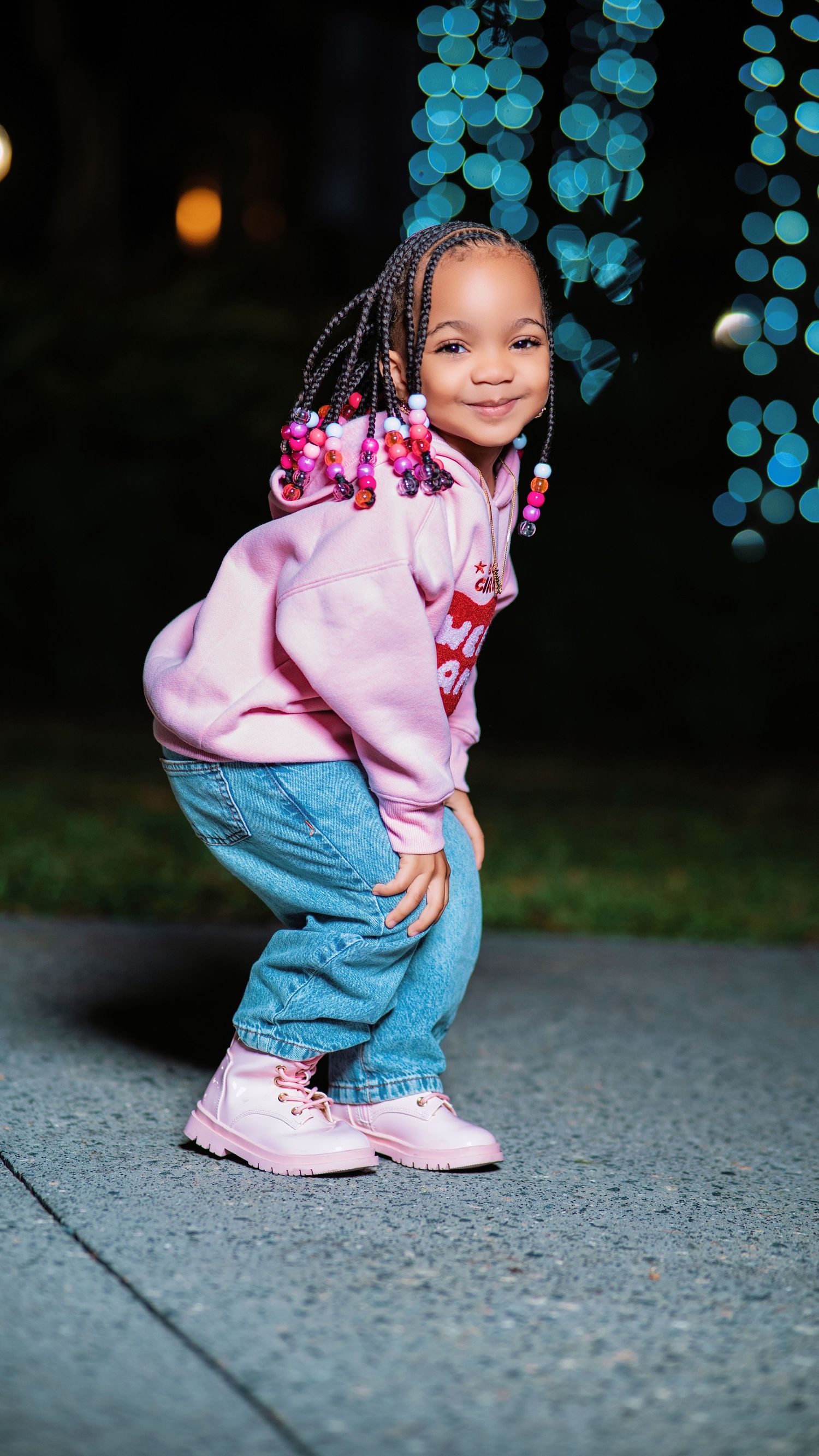 Small child in pink sweatshirt and denim poses against evening backdrop with blue lights.