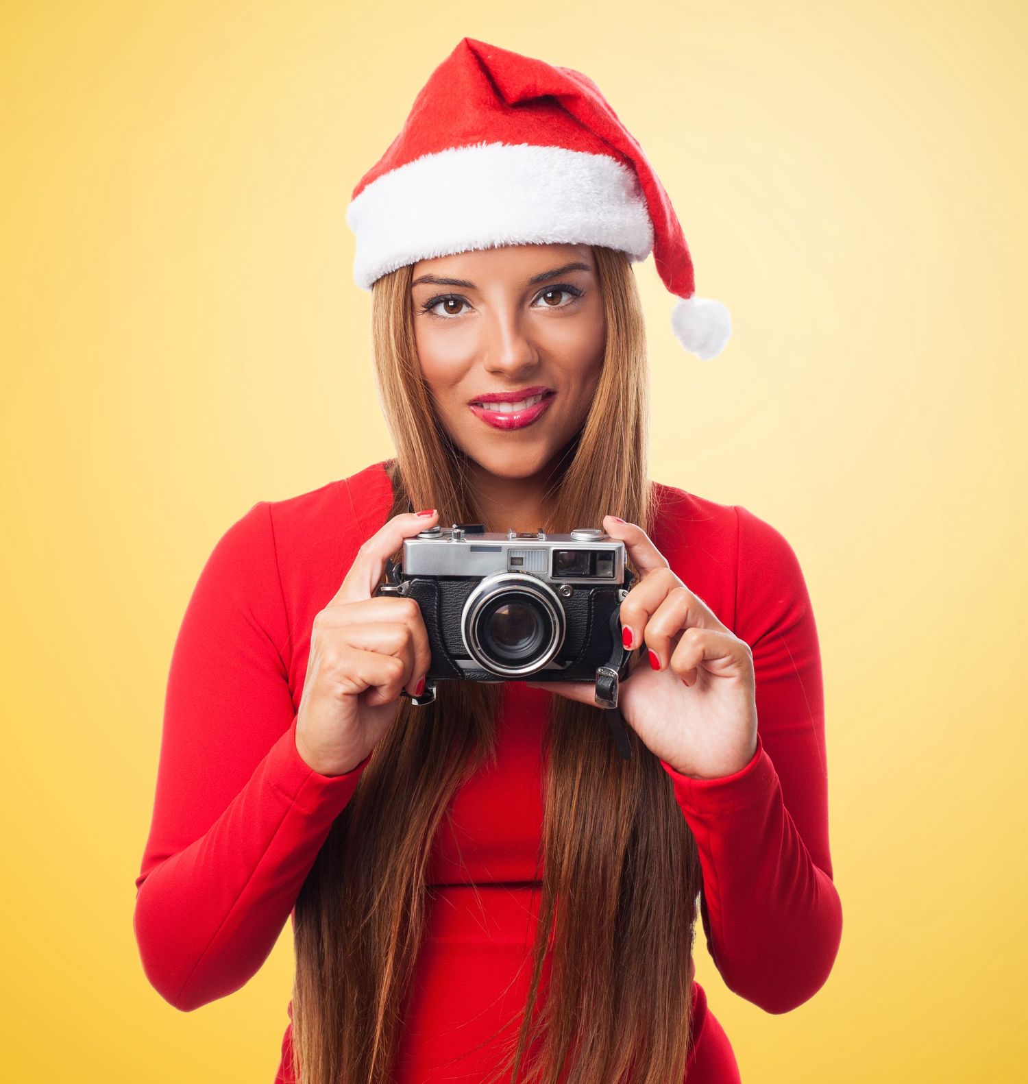 Person wearing red holiday outfit and Santa hat taking photos with vintage camera against yellow backdrop.