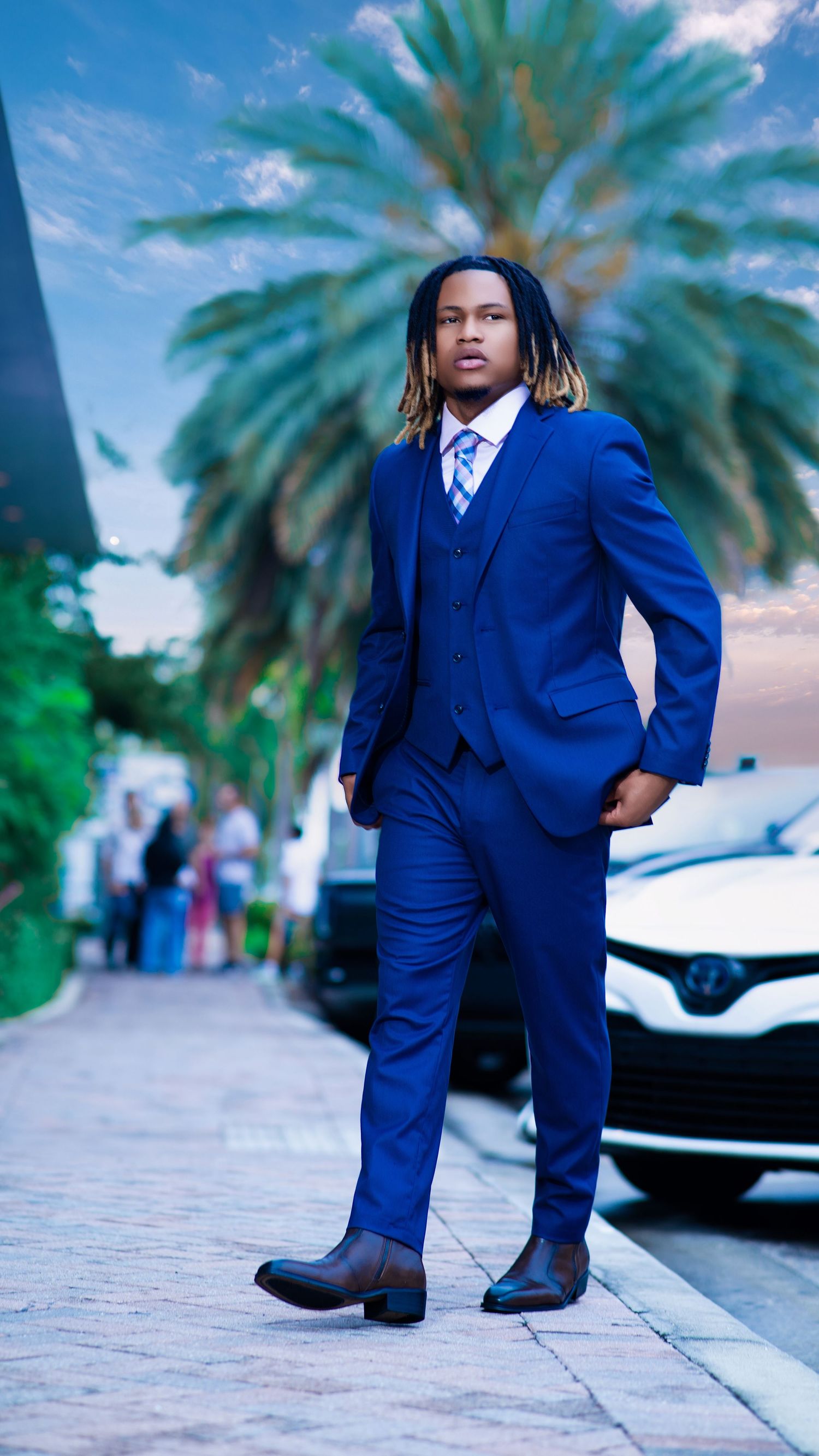 Young man in royal blue three-piece suit with tie, posing confidently outdoors near palm trees and parked cars.