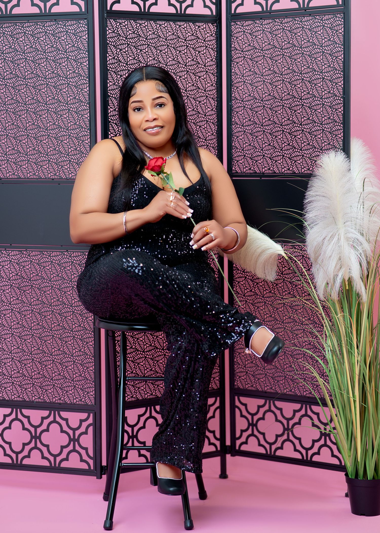 Woman in black sequin jumpsuit sits on stool holding red rose, posing in front of pink decorative screen.