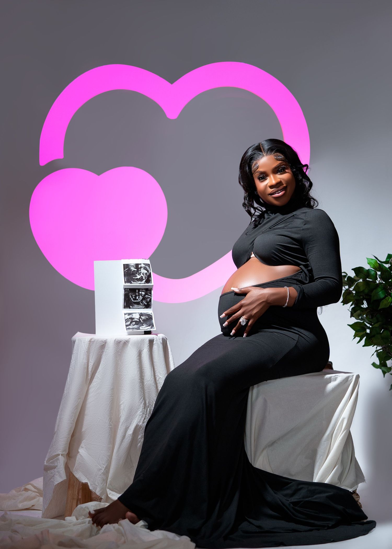 Pregnant woman in black dress poses for maternity photo with ultrasound images and pink heart backdrop.