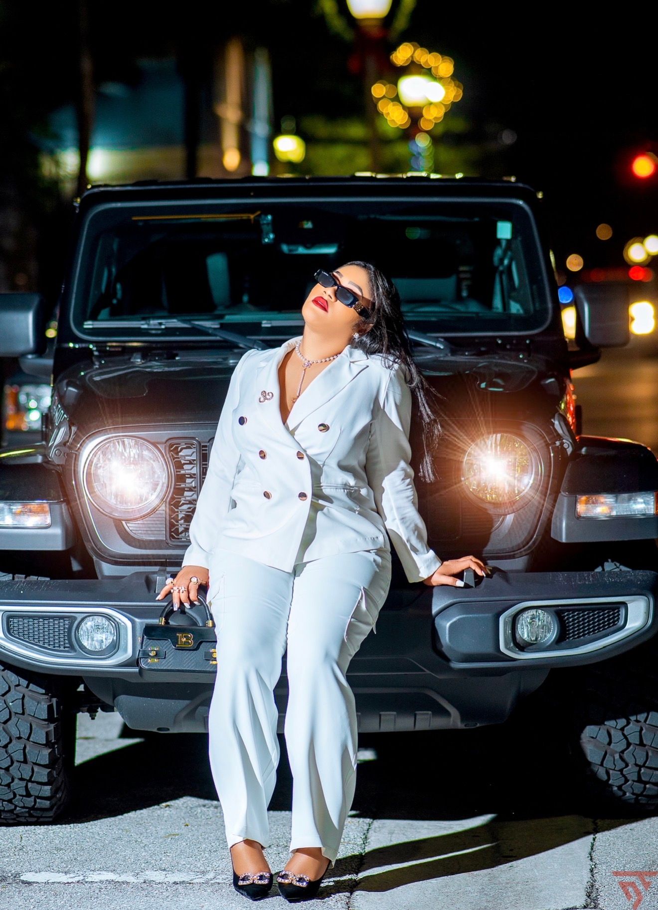 Woman in white pantsuit posing confidently in front of a black Jeep Wrangler at night with bokeh lights.