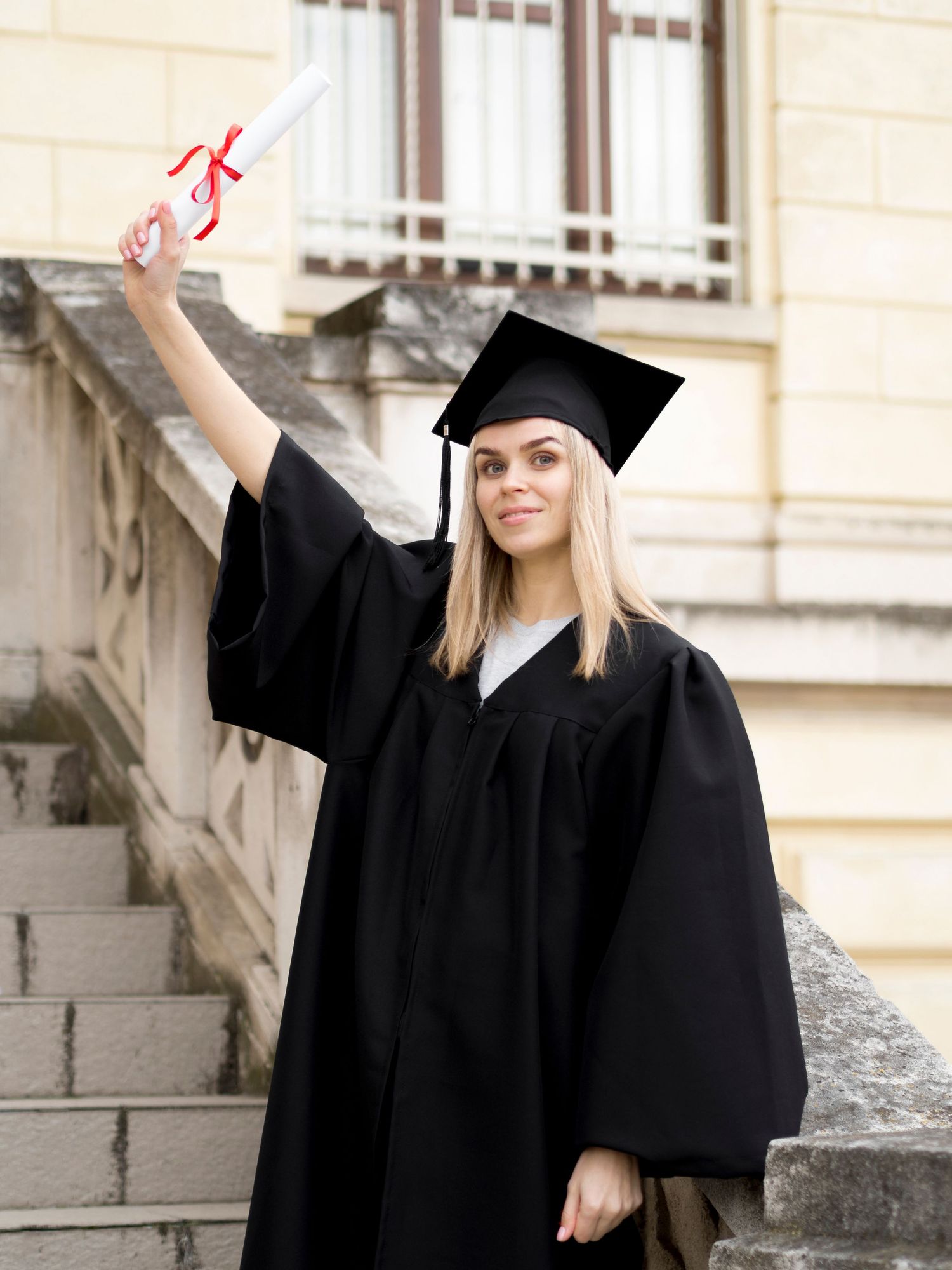 Graduate in black cap and gown celebrates with diploma on university steps.