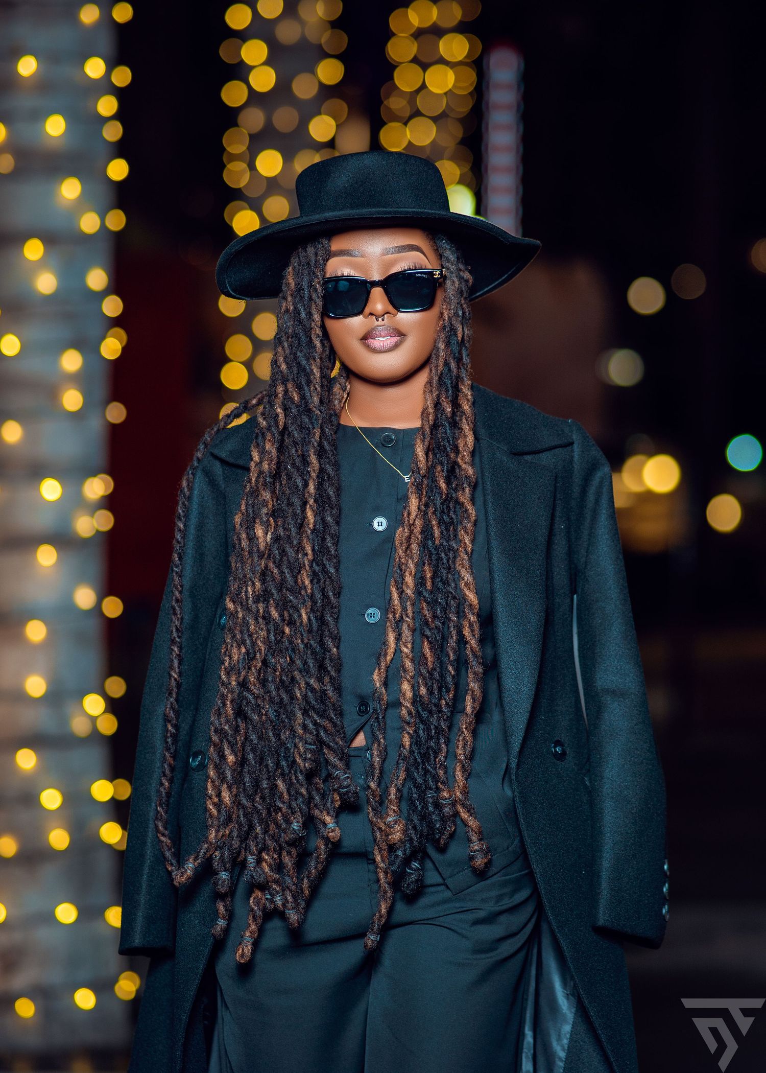 A stylish dark outfit with long braids and sunglasses against twinkling lights.