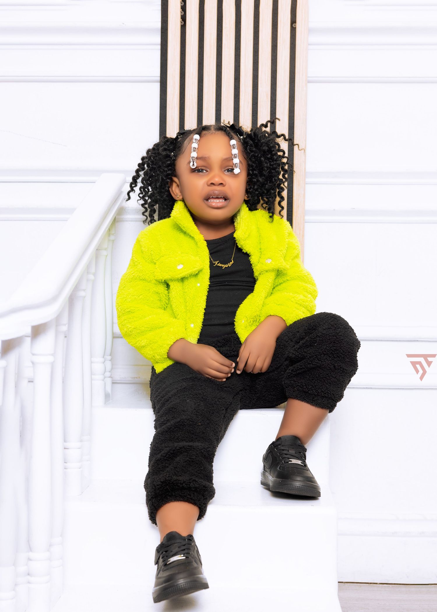 Child in neon yellow jacket and black outfit sits casually against white wall with vertical wood panels.