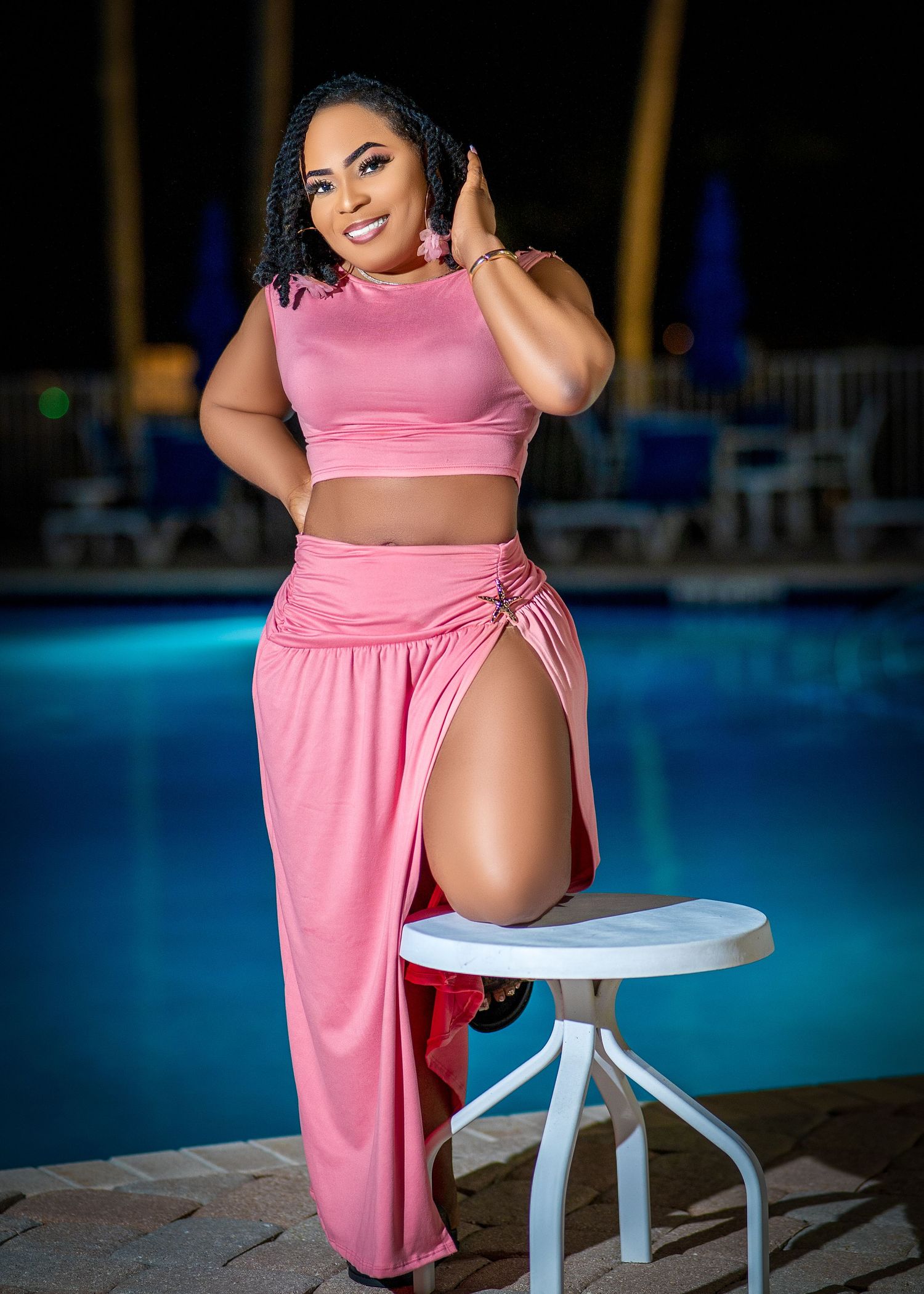 A person in a pink two-piece outfit poses confidently by a pool at night with a white table nearby.