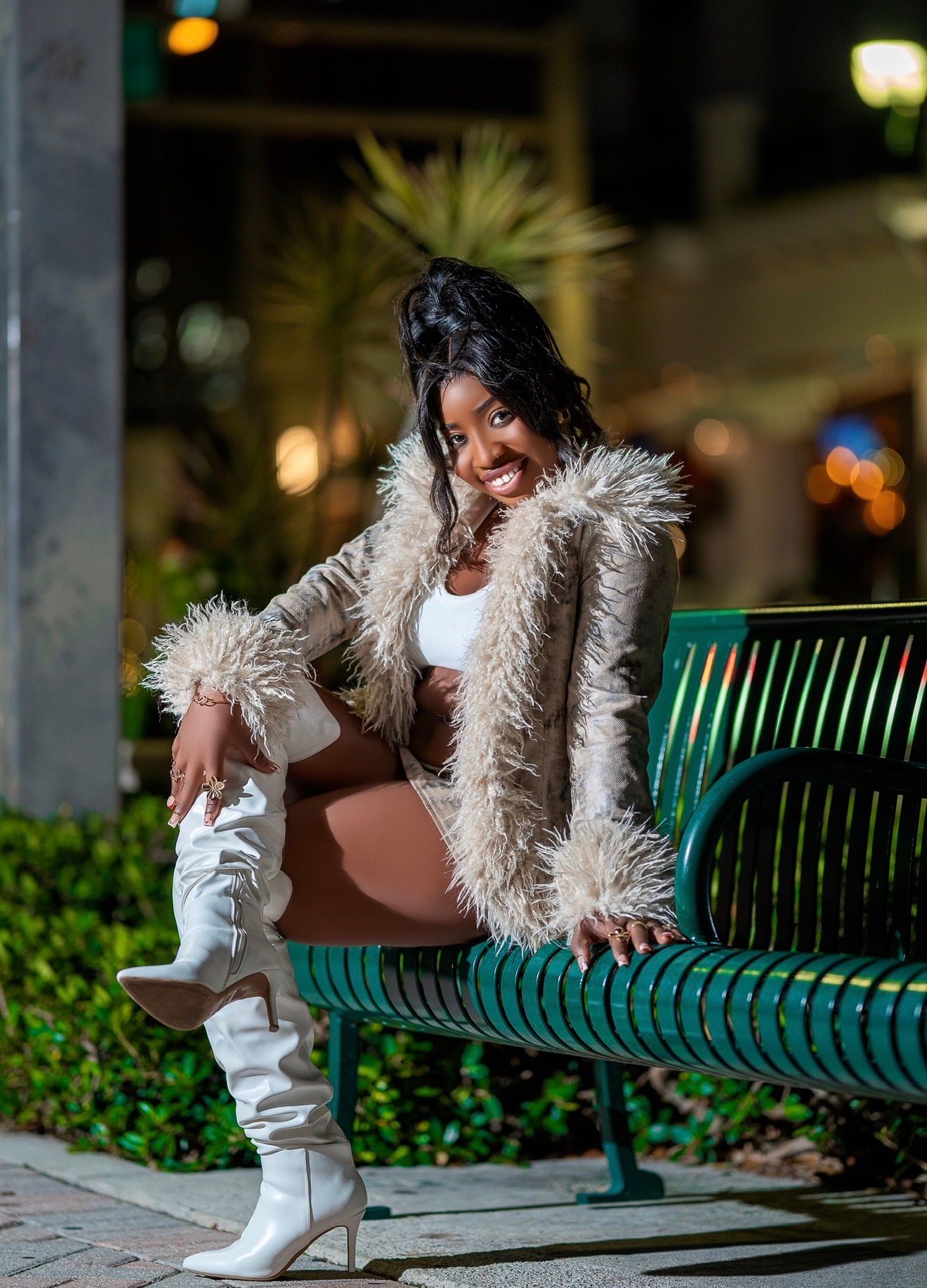 Smiling woman in fur coat and white knee-high boots sitting on a green bench outdoors at night.