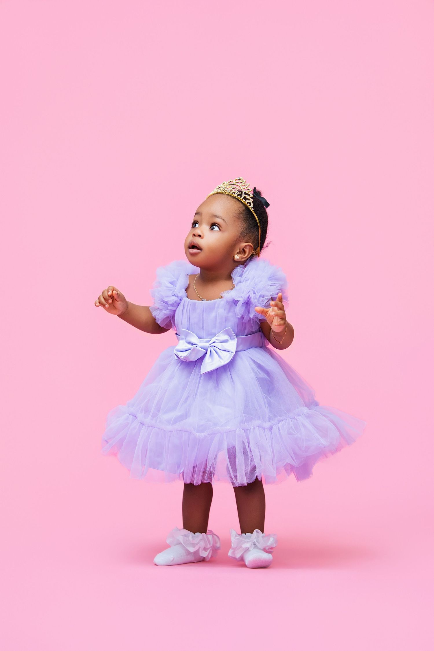 Adorable Black toddler girl in purple tutu dress and gold crown poses on pink background.