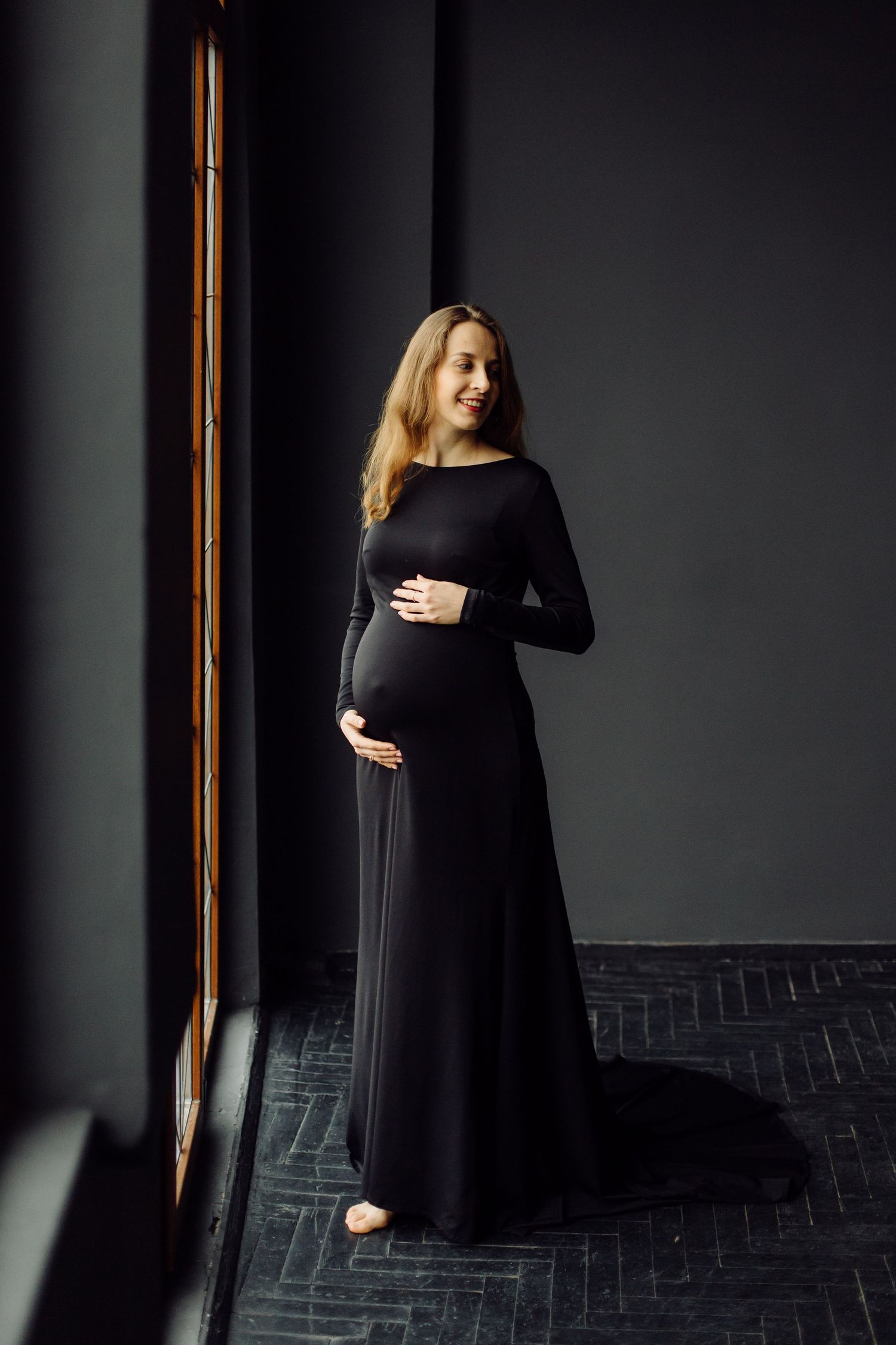 Moody maternity portraits in black dress against dark background with dramatic lighting.
