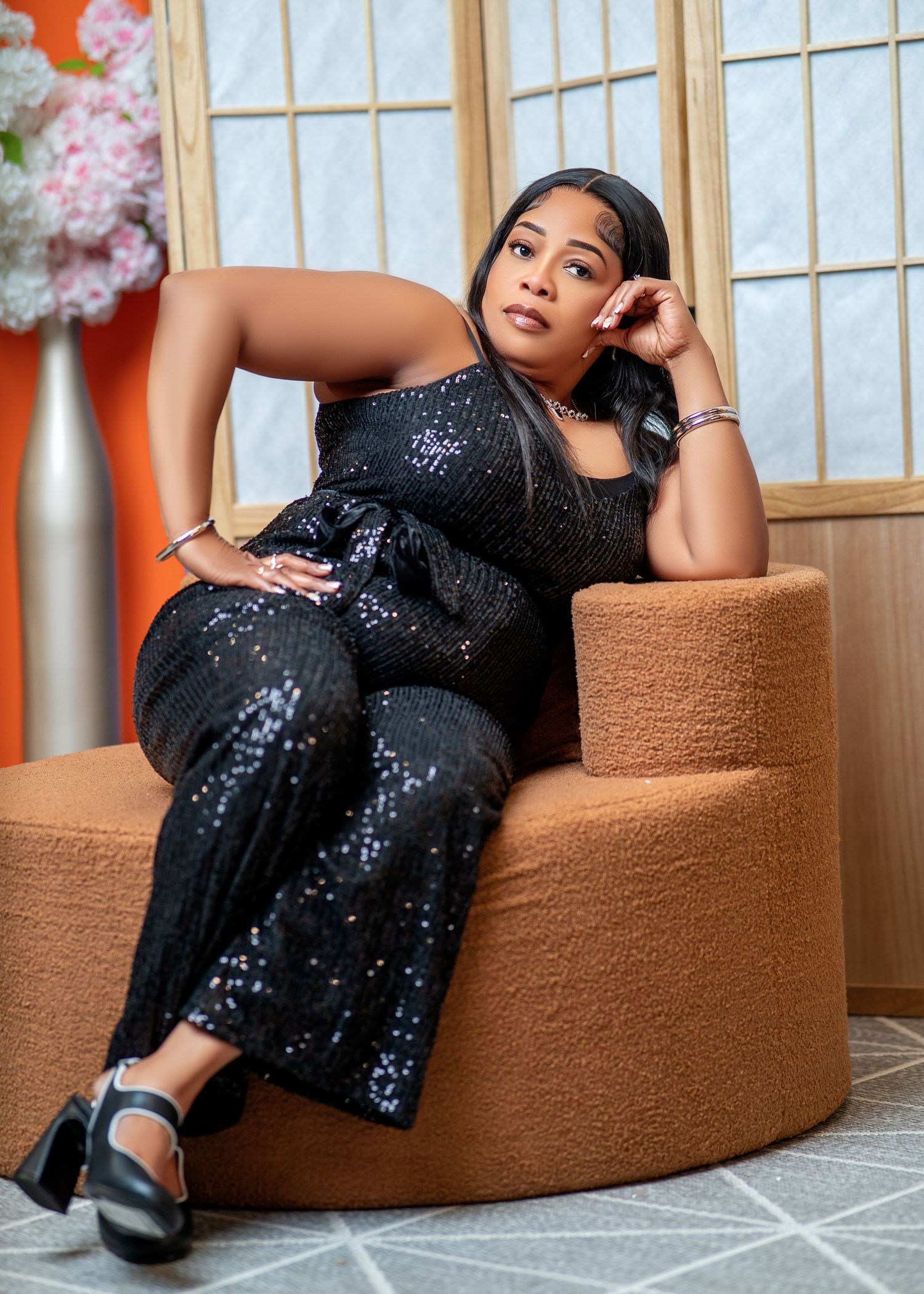 Woman in black sequin jumpsuit posing elegantly on a tan ottoman chair in a stylish orange-accented room.
