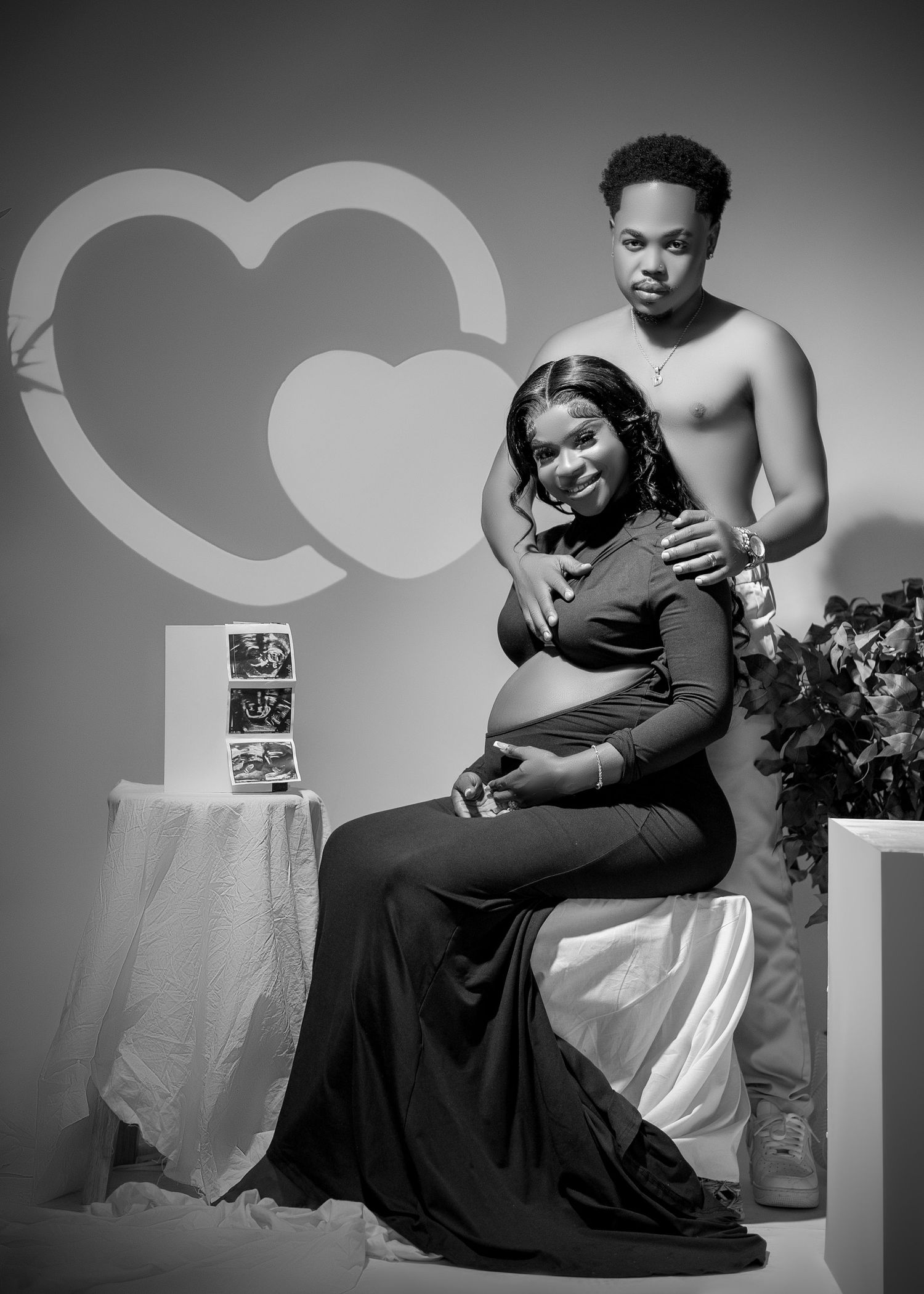Black and white maternity photo of a pregnant woman in a gown with her partner, hearts backdrop, ultrasound photos displayed.