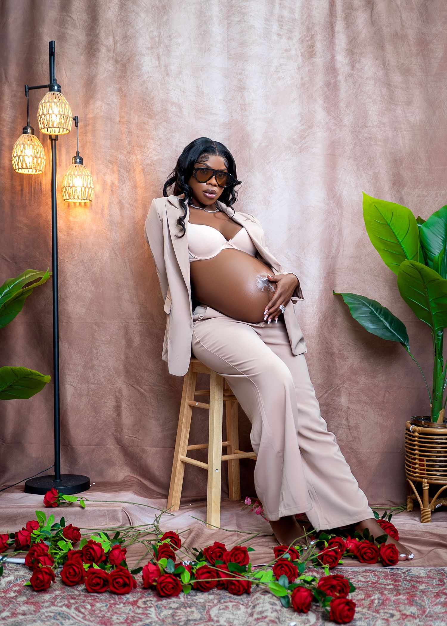 Pregnant woman in beige blazer and white bra poses on stool with red roses, plants, and warm lamp in studio.