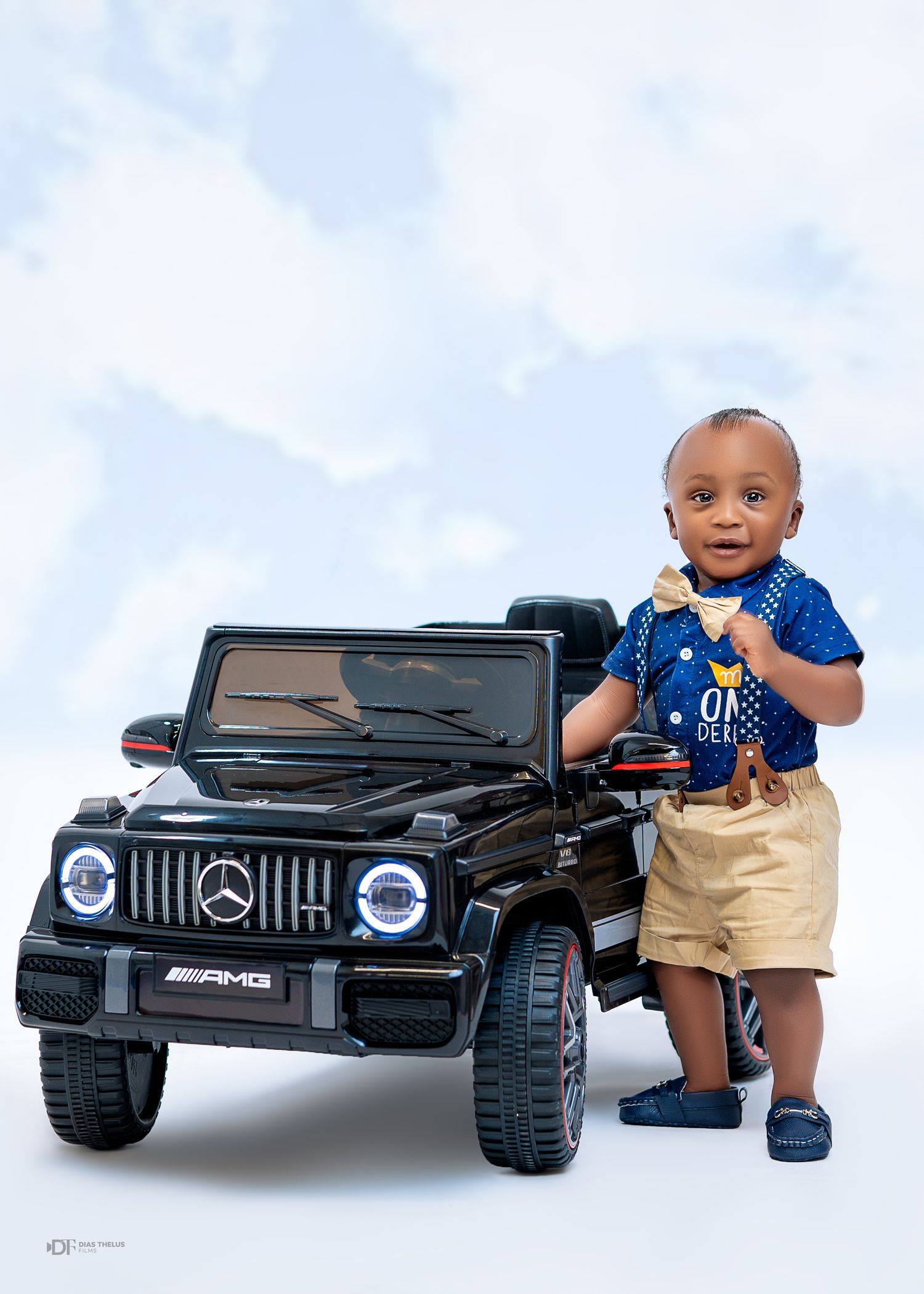 Toddler boy in blue shirt and khaki shorts standing next to a black Mercedes-Benz AMG G-Class ride-on toy car.