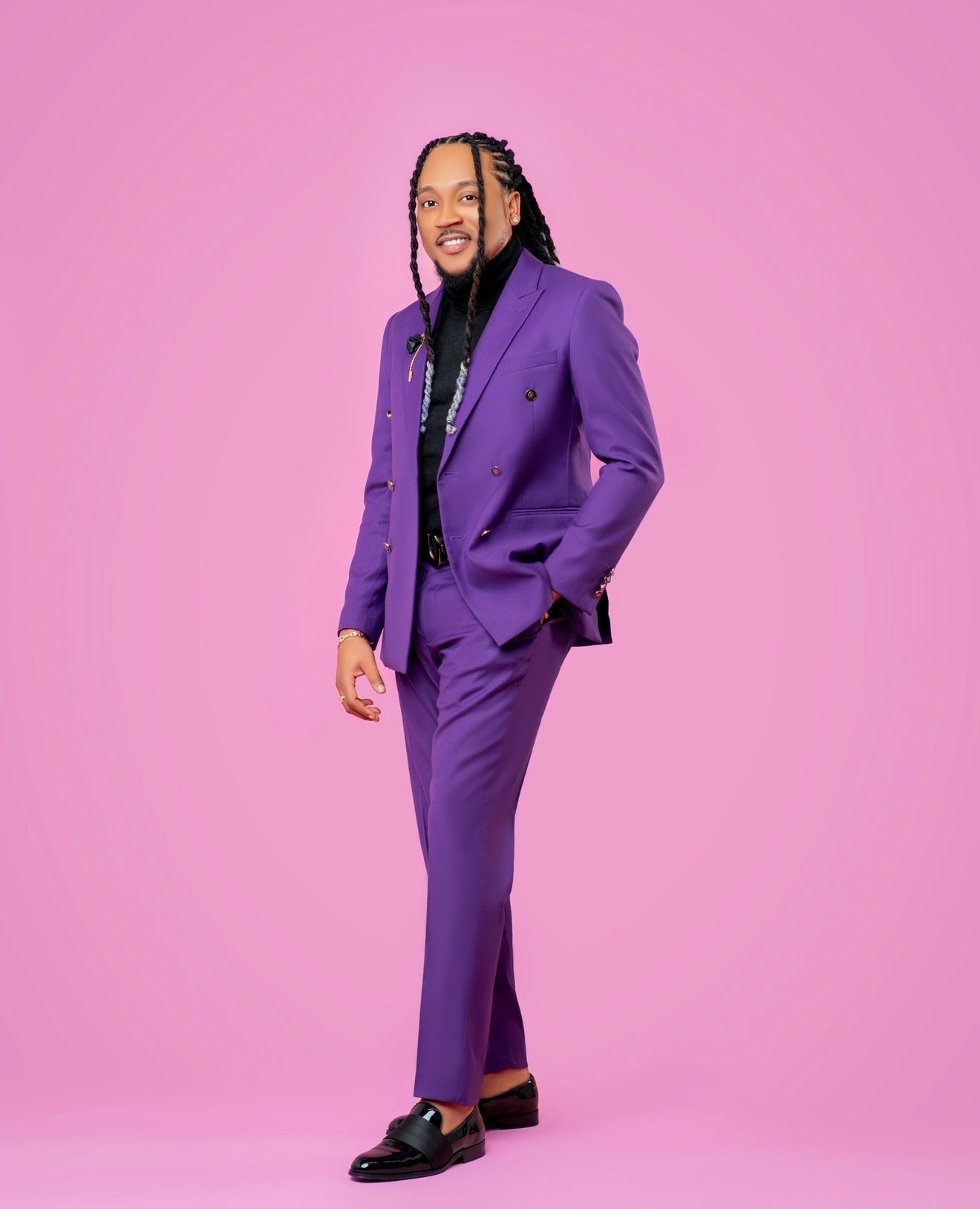 A stylish man wearing a bold purple double-breasted suit with braided hair poses confidently against a pink background.