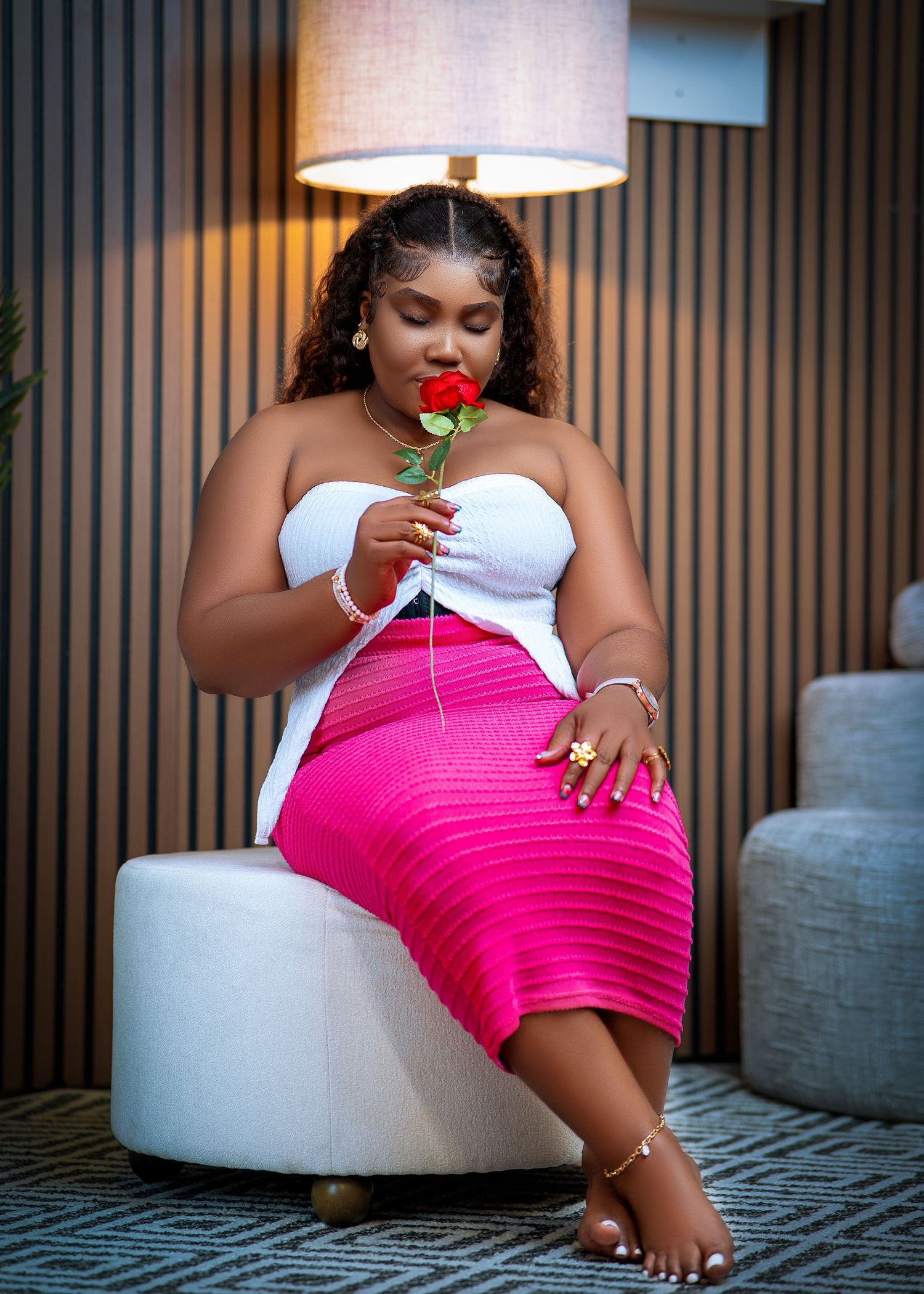 Woman in white top and pink skirt sitting on chair, smelling a red rose near a lamp.