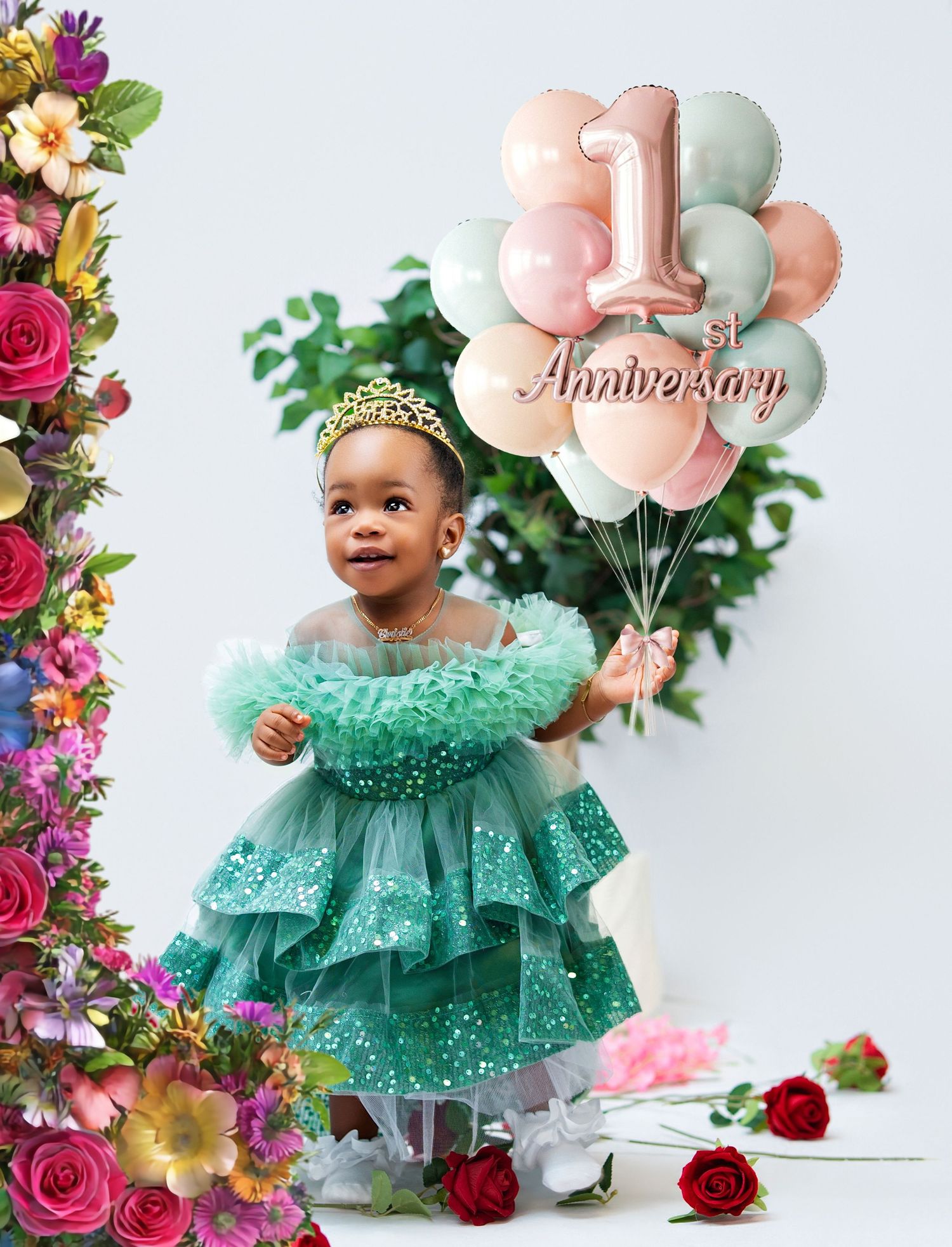 Toddler girl in green tulle dress with gold tiara holds 1st Anniversary balloons surrounded by colorful flowers.