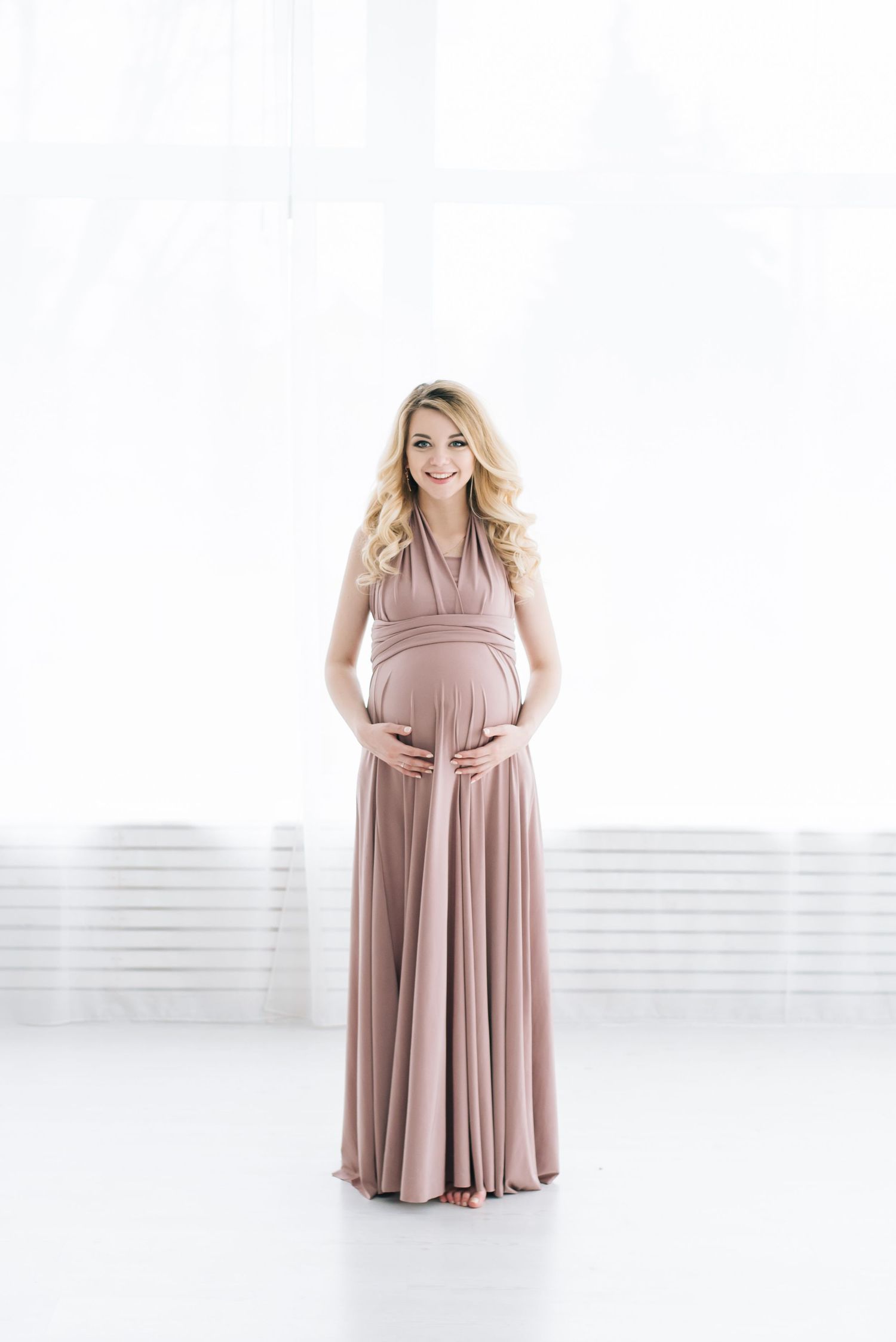 Elegant maternity photos in a flowing dusty rose dress against white backdrop.