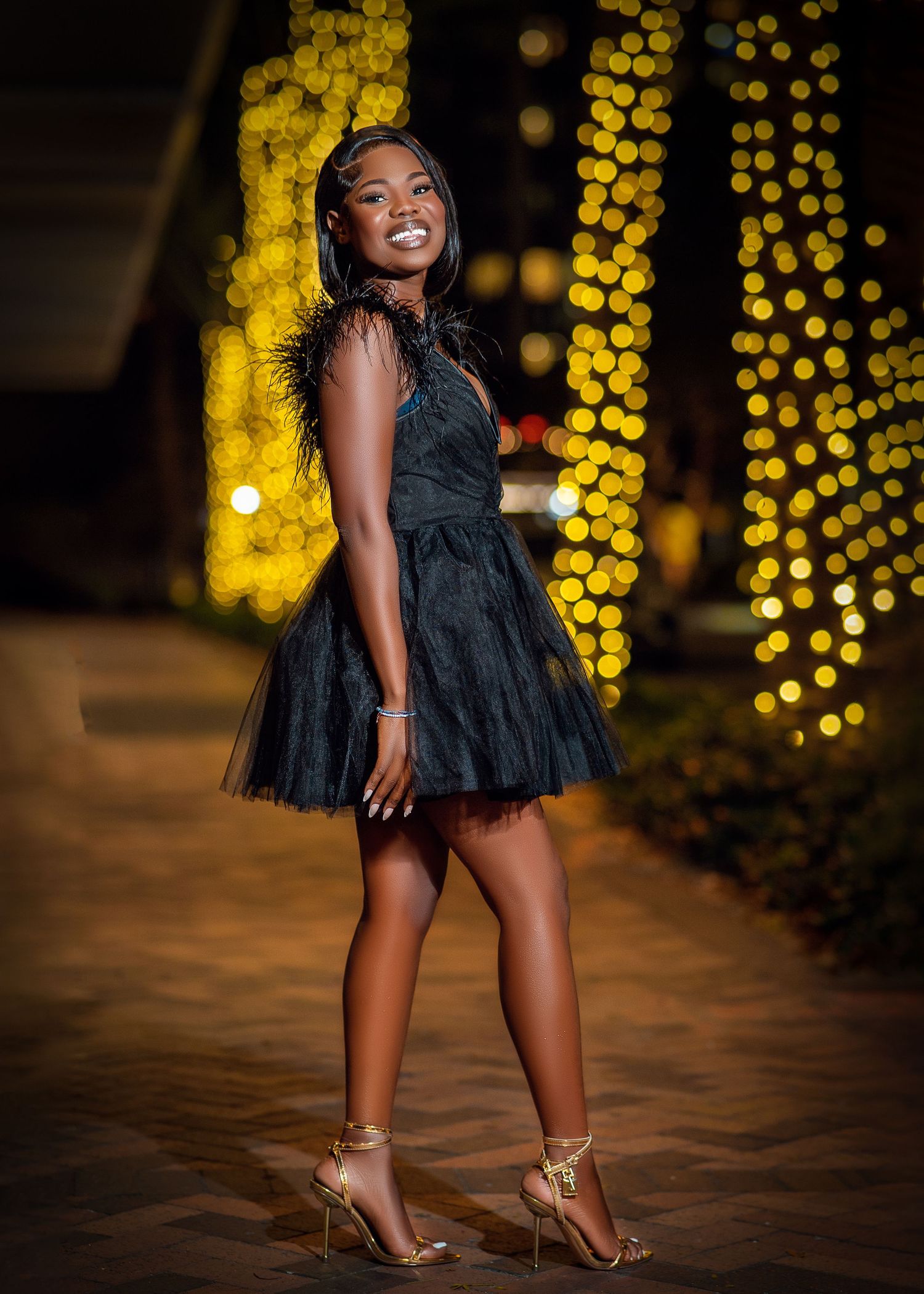 A person in a black dress poses playfully against a backdrop of glowing yellow bokeh lights and wooden flooring.