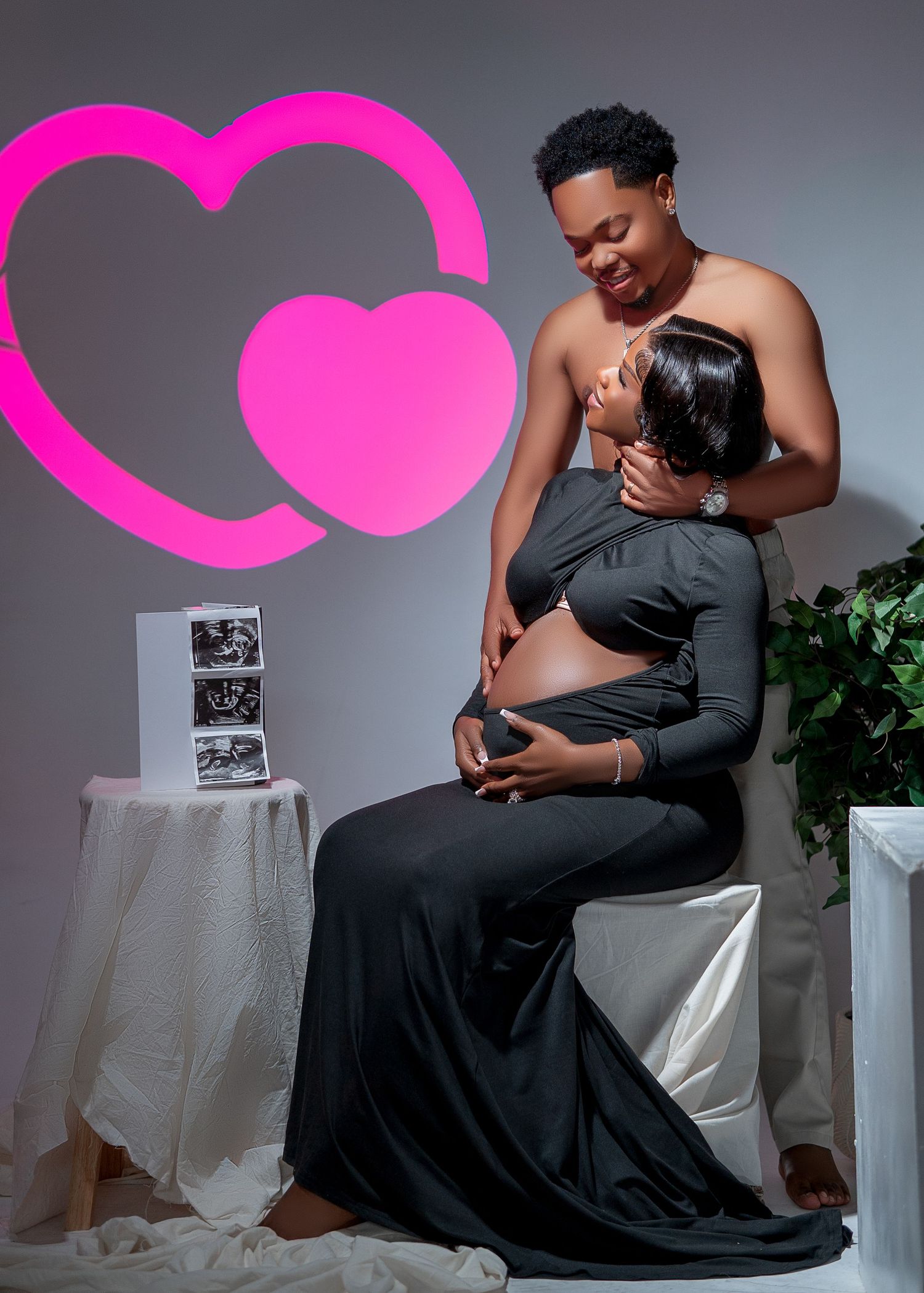 Couple maternity photo shoot in black outfit with pink hearts and ultrasound pictures in a studio setting.